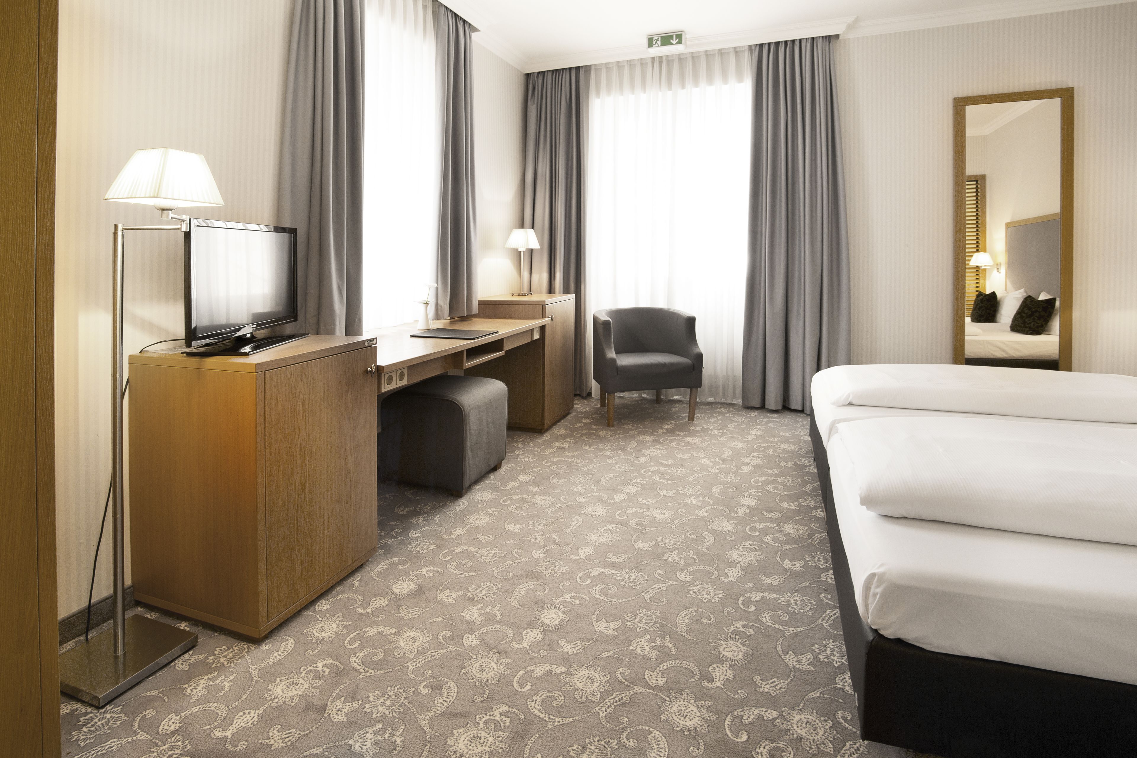 A hotel room with a bed, a desk with a computer, a chair, a TV, and a large mirror.