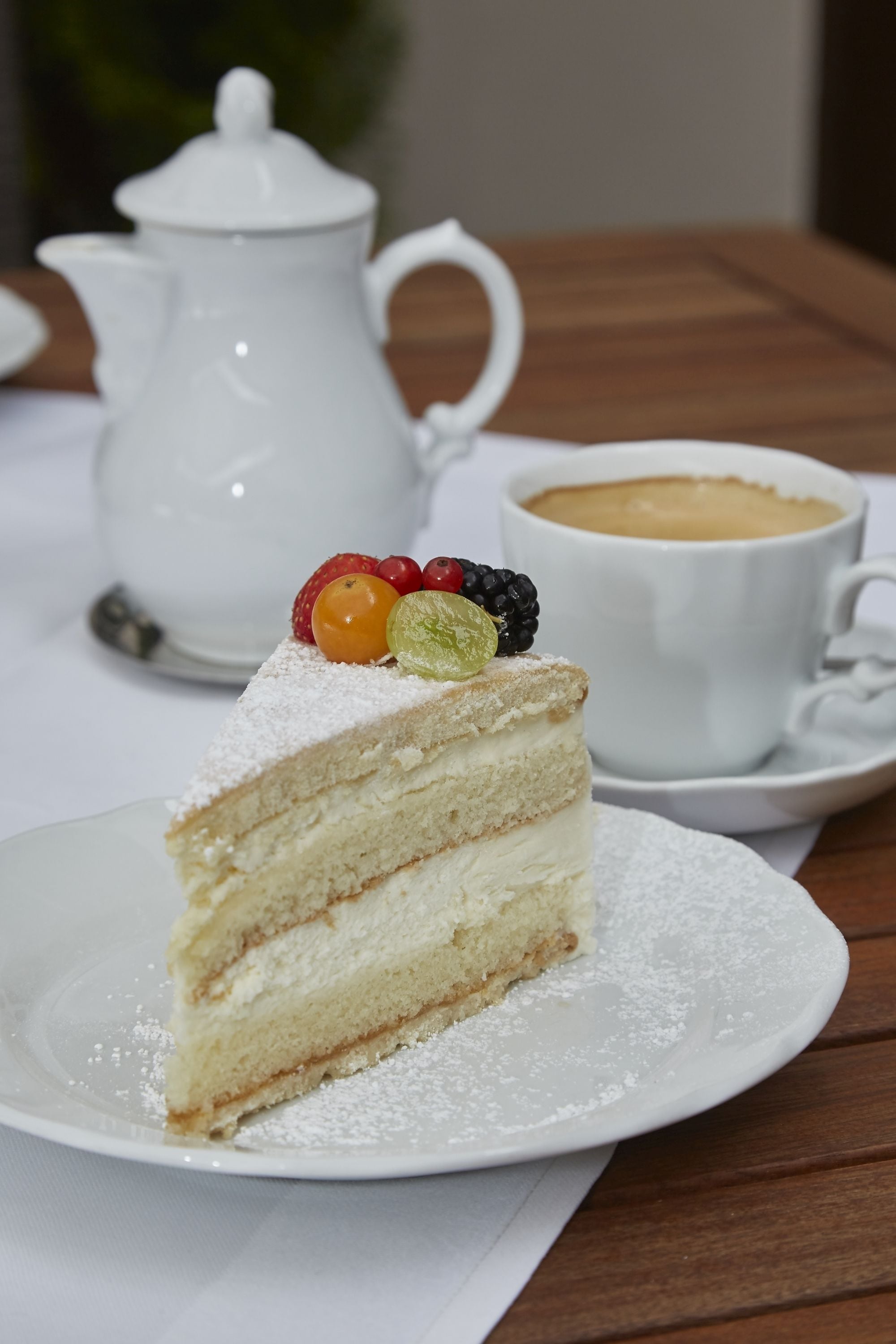 Une part de gâteau à plusieurs couches, garnie de fruits, est posée sur une assiette. À côté, on aperçoit une théière blanche et une tasse de café sur une table en bois.