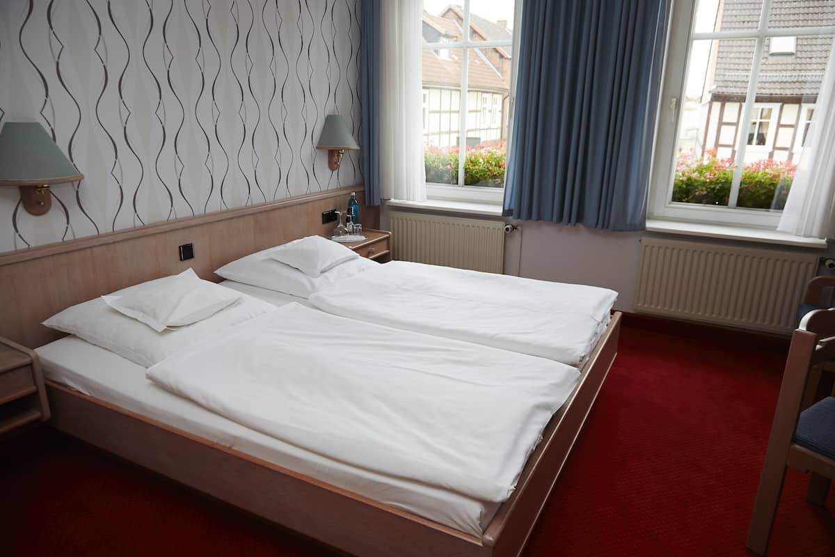 Un lit double avec du linge de lit blanc, une table de chevet avec une lampe, et une vue sur un immeuble à travers une fenêtre ornée de rideaux bleus.