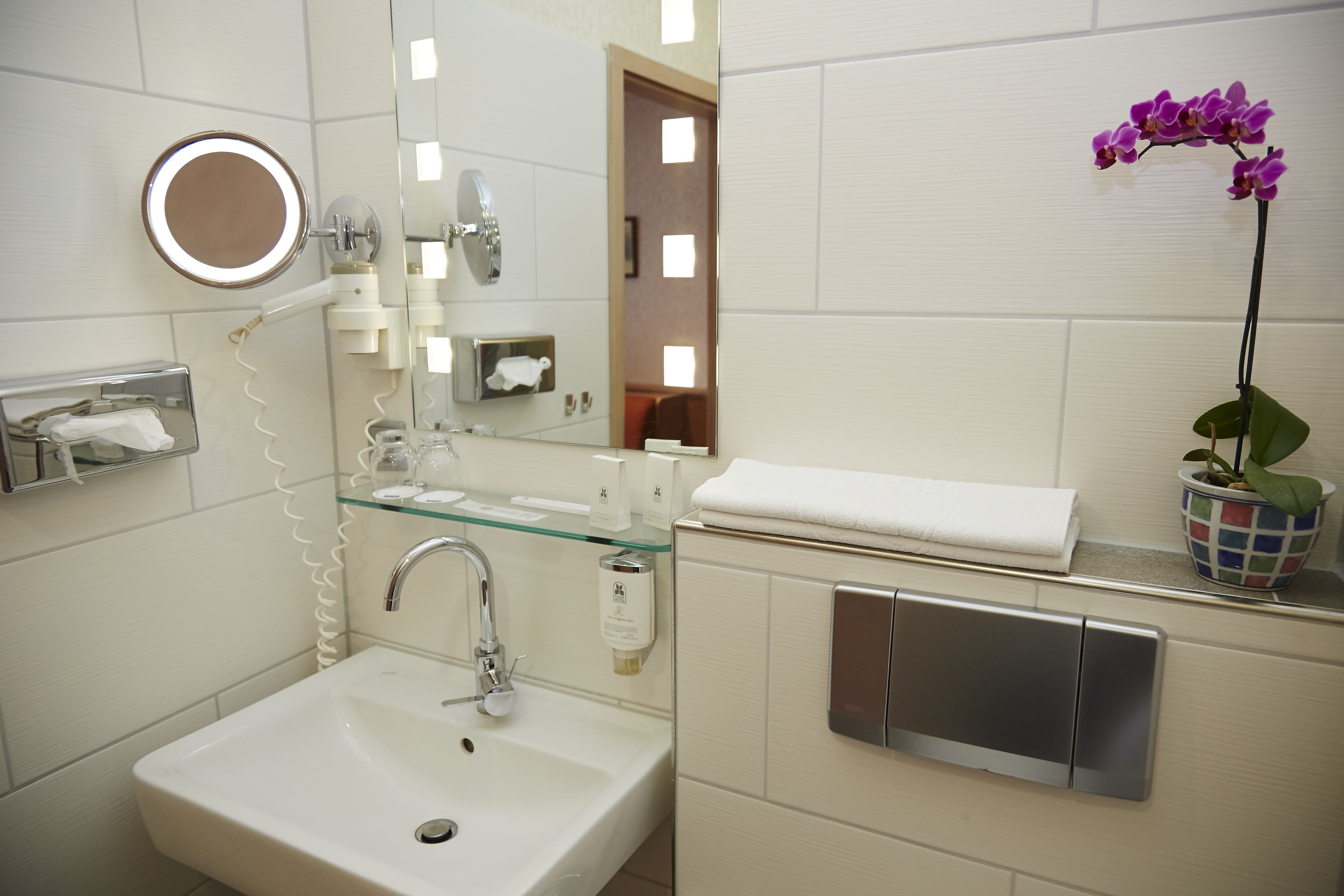 A bathroom with a white sink, a glass shelf with toiletries, a mirror with a light, and a potted orchid.