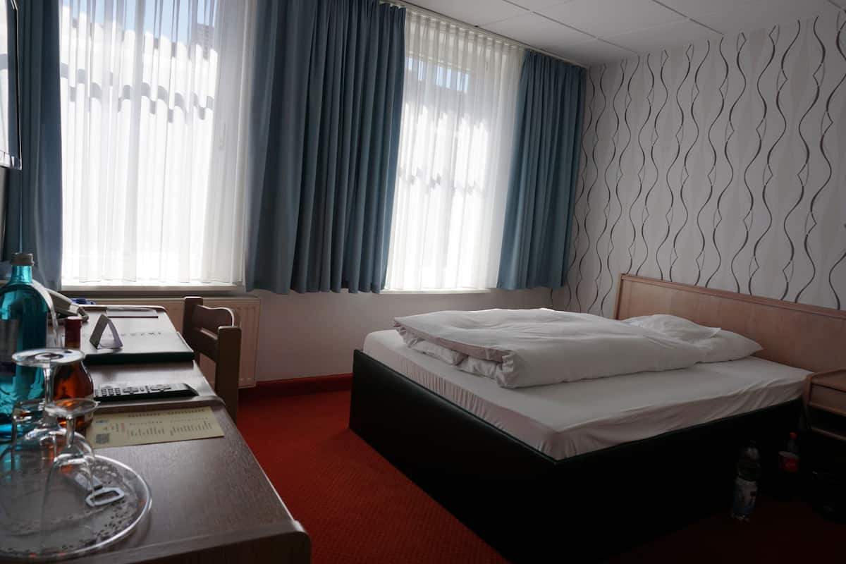 Une chambre d’hôtel avec un lit, un bureau avec un ordinateur portable, une chaise et une fenêtre avec des rideaux.