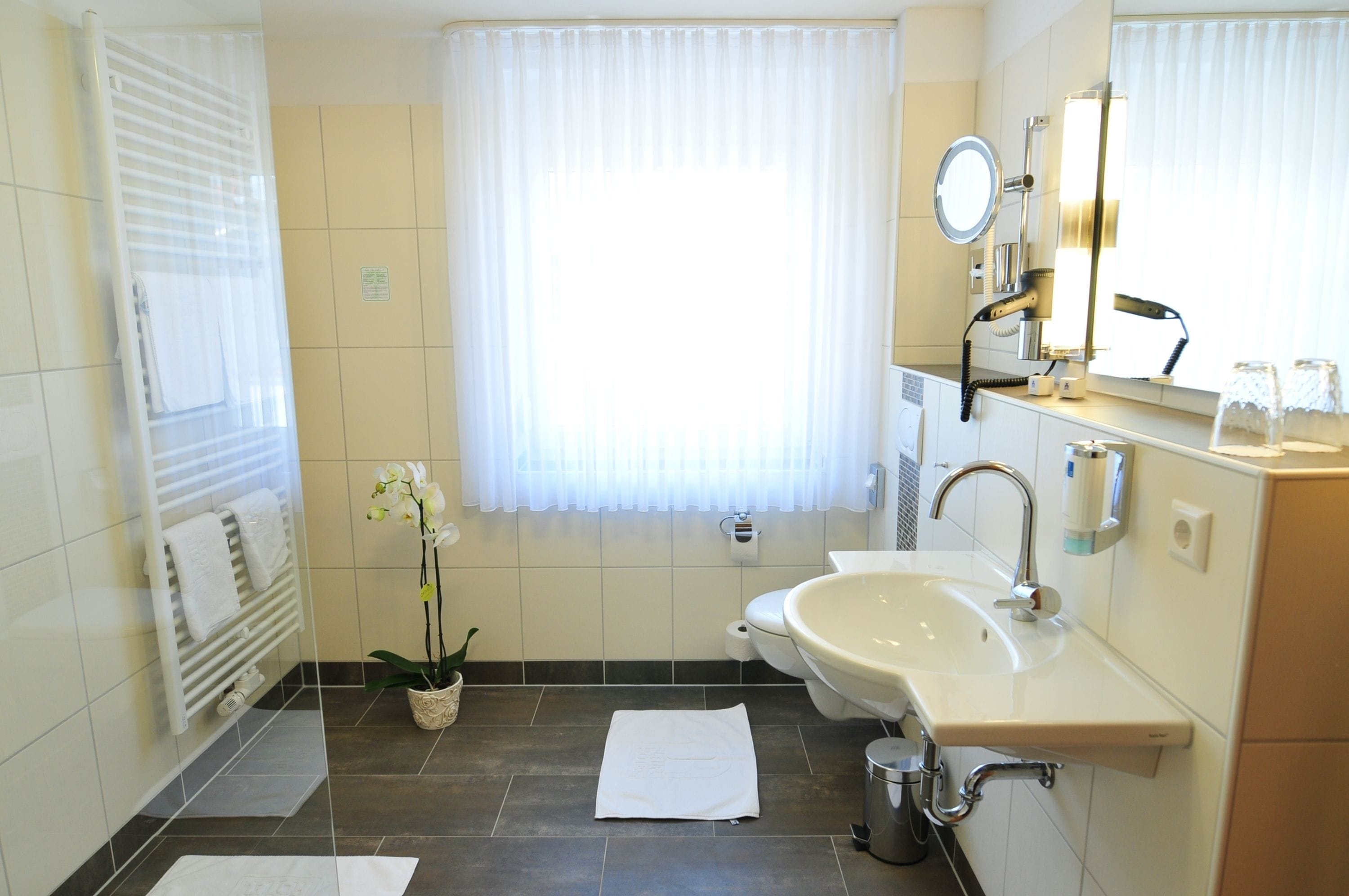 A modern bathroom with a bathtub, sink, and toilet. There is a window with sheer curtains, a towel rack, and a potted orchid.