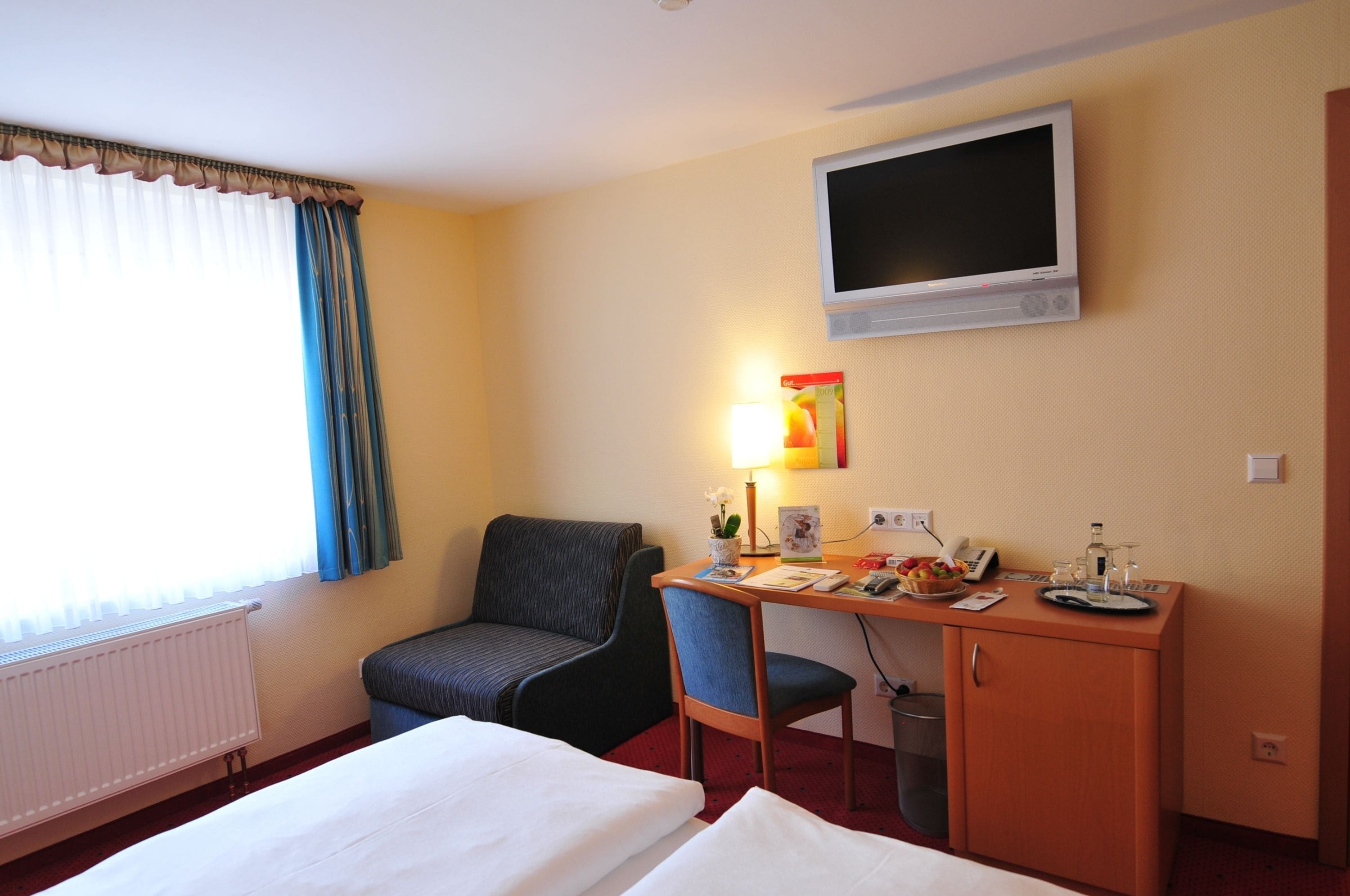 A hotel room with a bed, a desk with a lamp, a chair, a television mounted on the wall, and a radiator.