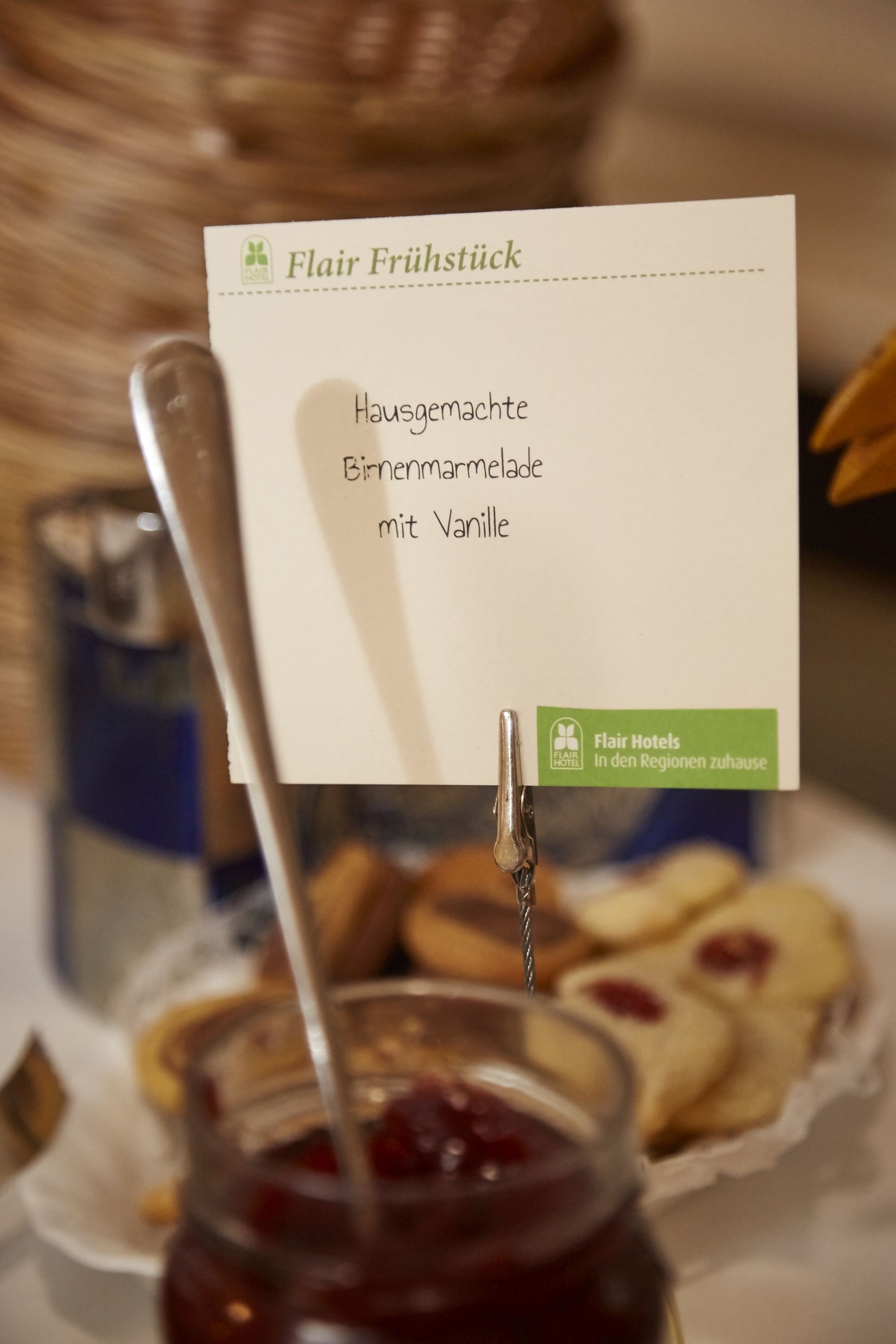 Tarjeta de menú titulada 'Flair Frühstück' con opciones como 'Mermelada casera de peras con vainilla' y un tarro de mermelada.