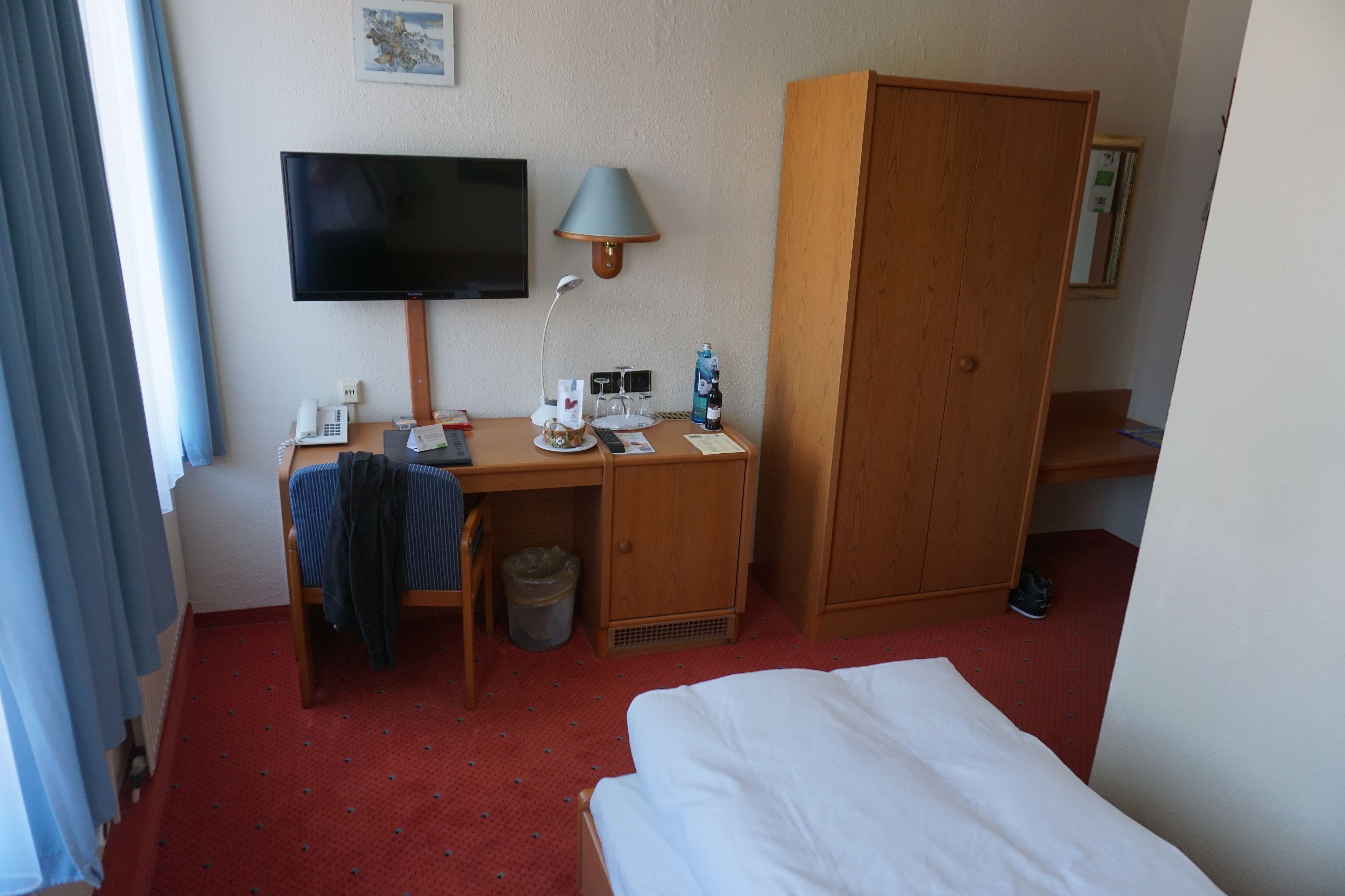 Una habitación de hotel con cama, escritorio con computadora, televisor, lámpara y armario.