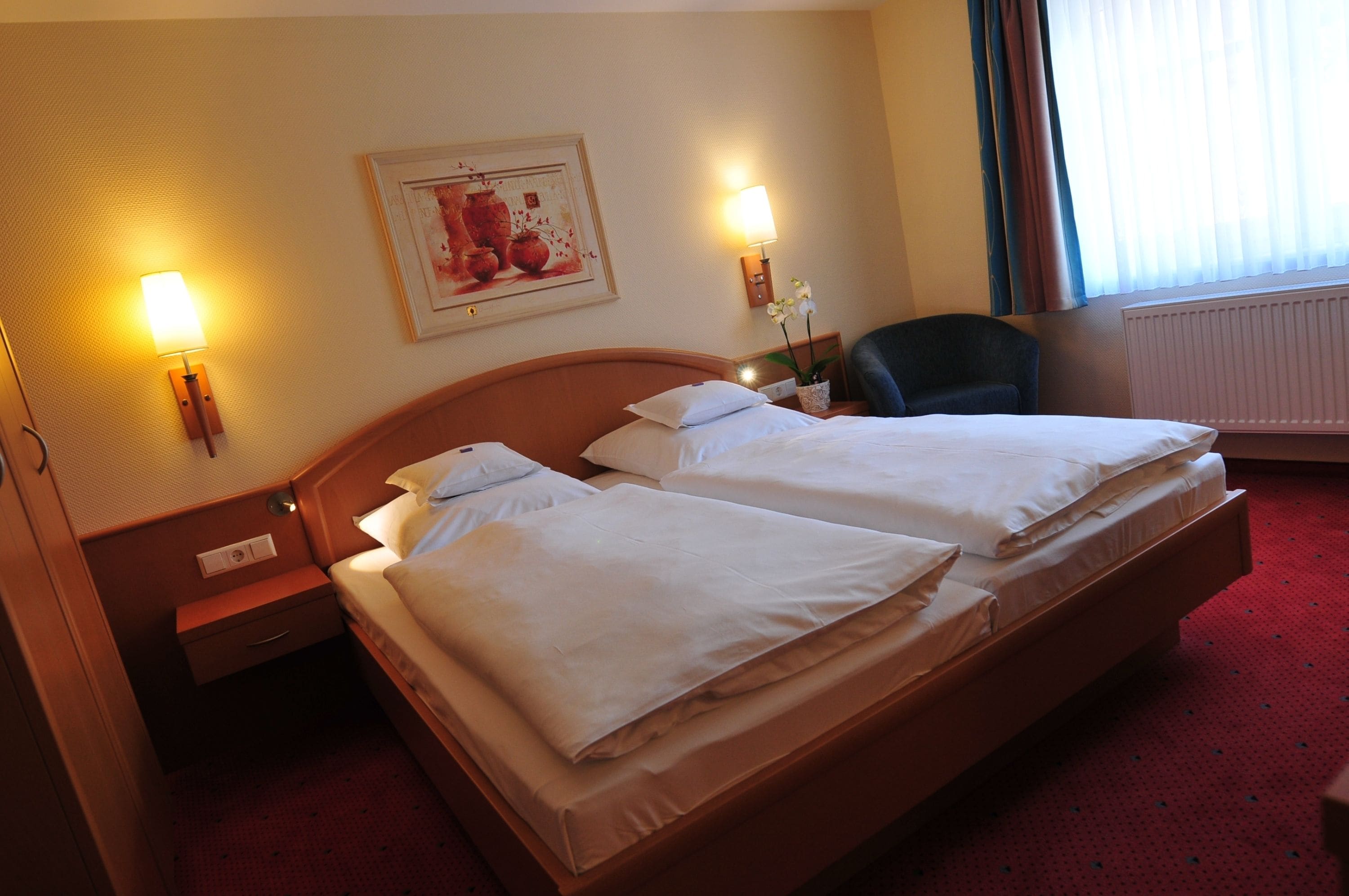 A hotel room with two beds, a nightstand, a lamp, a chair, and a painting on the wall.