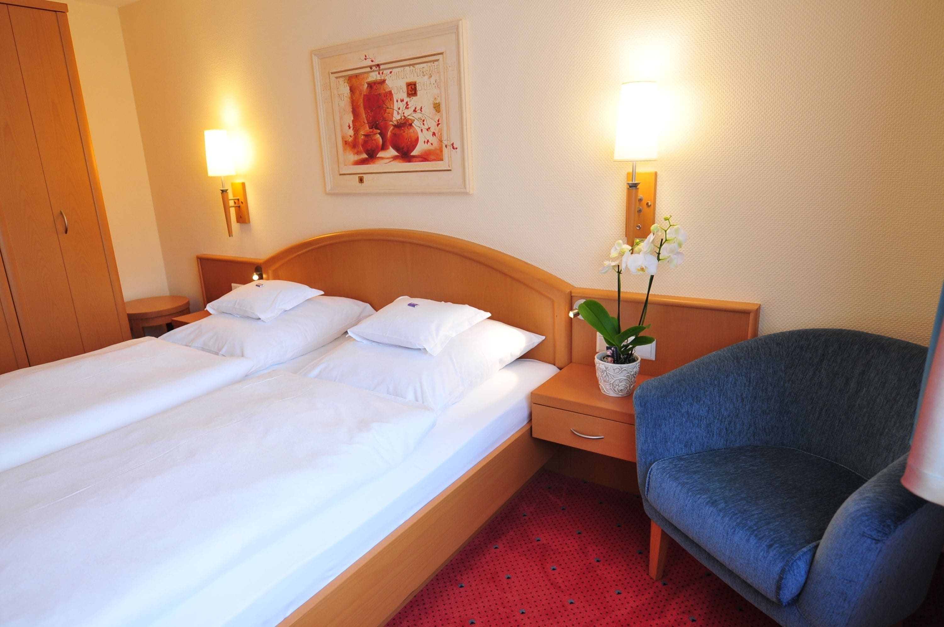A hotel room with a double bed, a nightstand with a lamp, a chair, and a wall-mounted artwork.