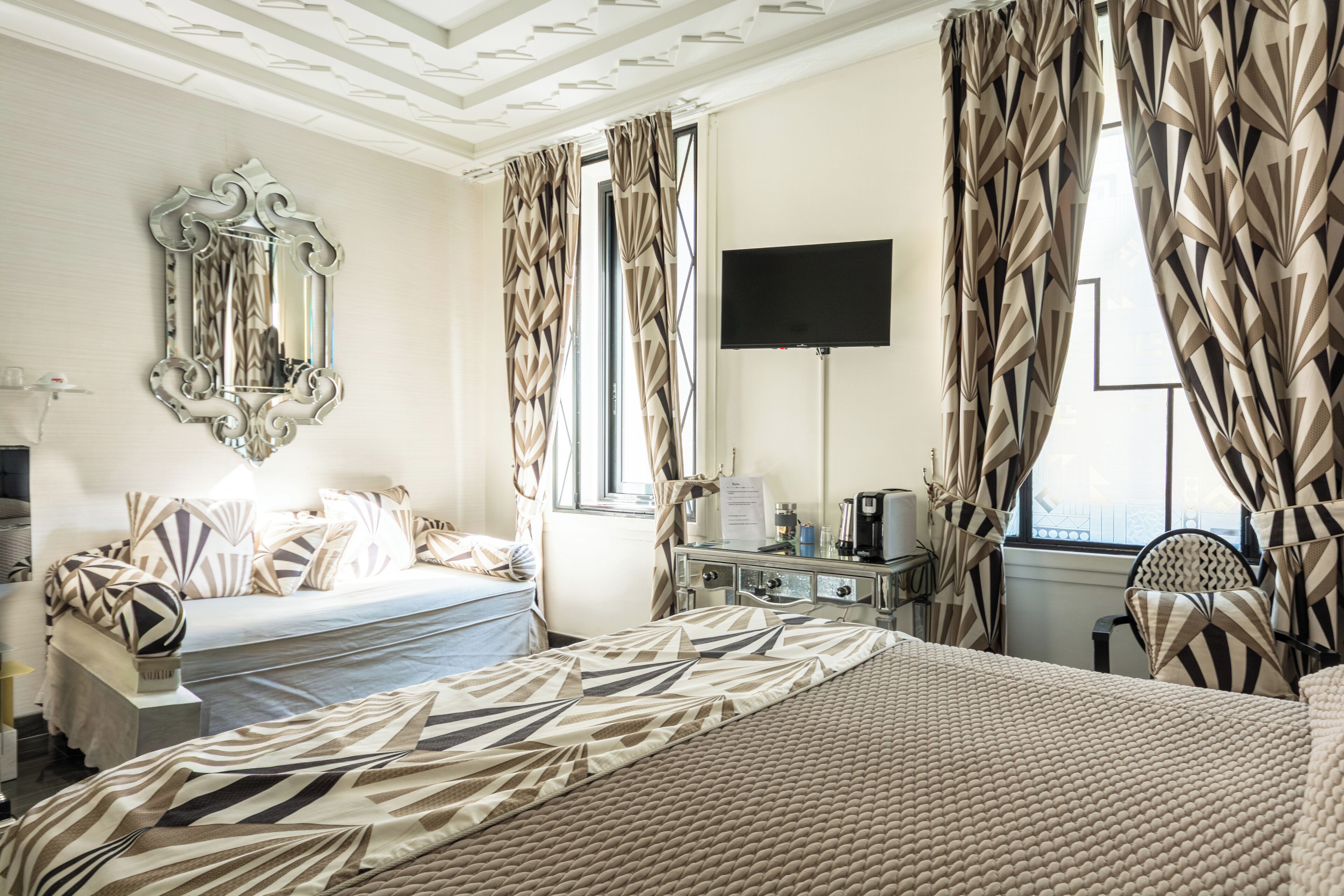 A hotel room with a bed, a desk with a coffee maker, a TV, and curtains with a geometric pattern.