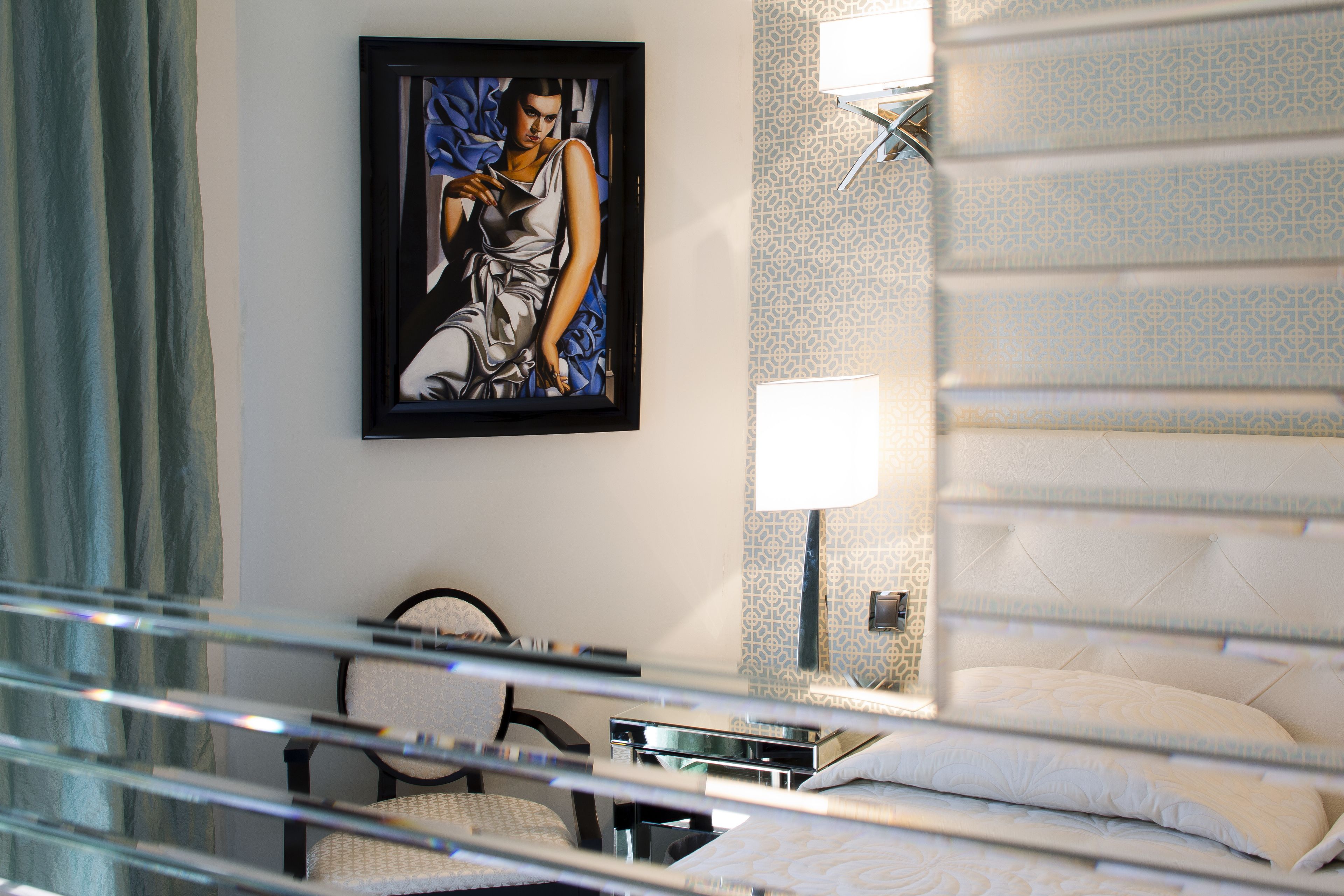 A hotel room with a bed, a chair, a desk, a framed artwork on the wall, and a patterned wall.