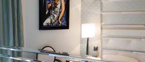 A hotel room with a bed, a chair, a desk, a framed artwork on the wall, and a patterned wall.