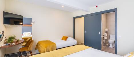 Quarto de hotel com cama, duas mesas de cabeceira, escrivaninha com cadeira, televisão e banheiro visível por uma porta aberta.