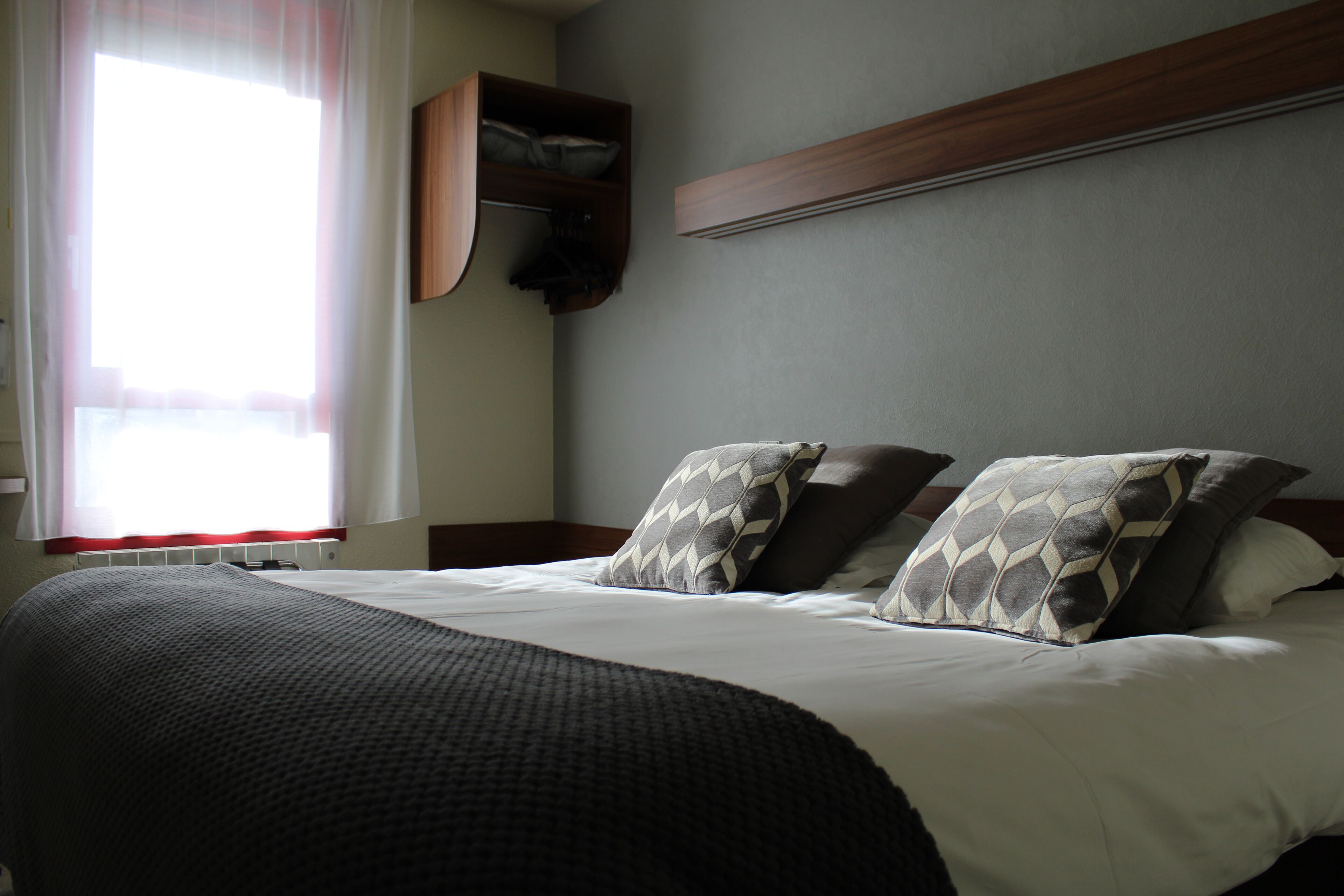 A neatly made bed with a dark blanket and patterned pillows, a wooden headboard, and a built-in shelf above the bed.