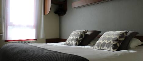 A neatly made bed with a dark blanket and patterned pillows, a wooden headboard, and a built-in shelf above the bed.