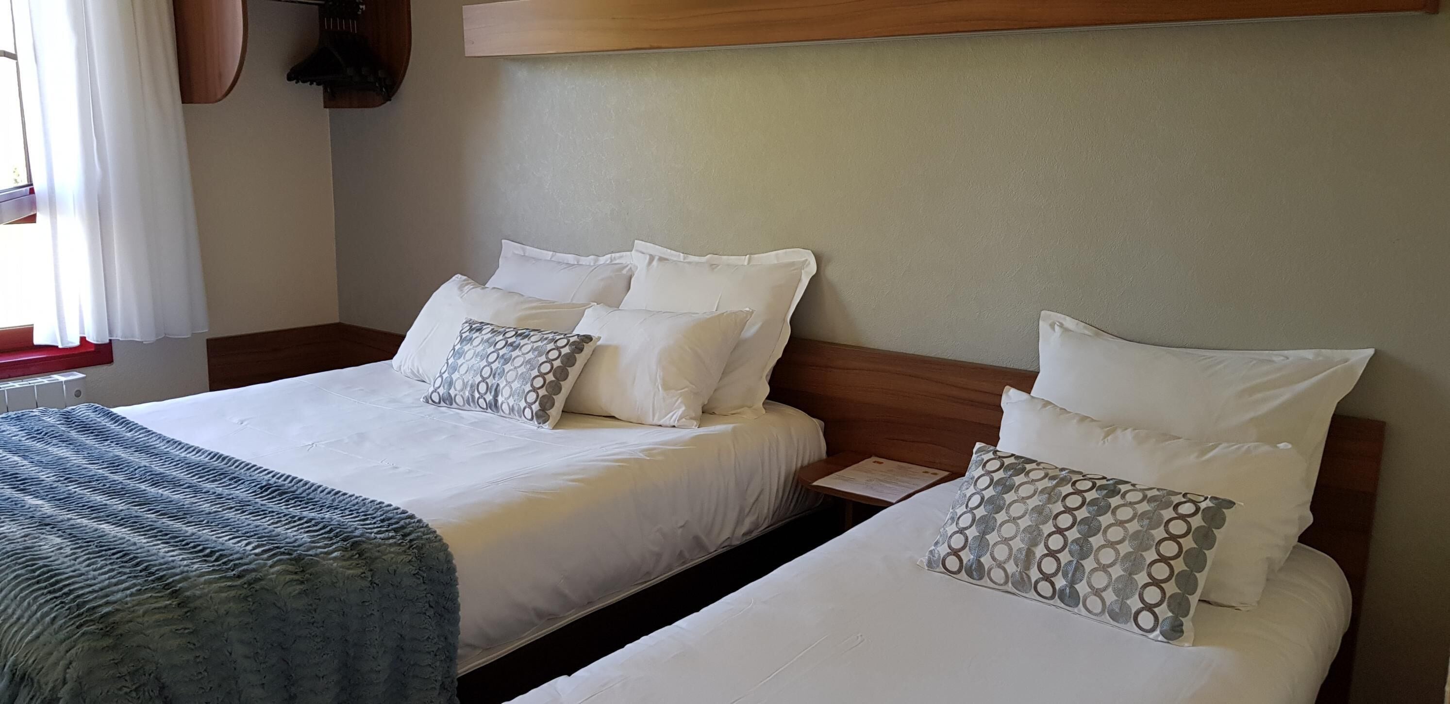 A hotel room with two beds, each with white bedding and patterned pillows. There is a wooden headboard, a small table with a menu, and a window with curtains.