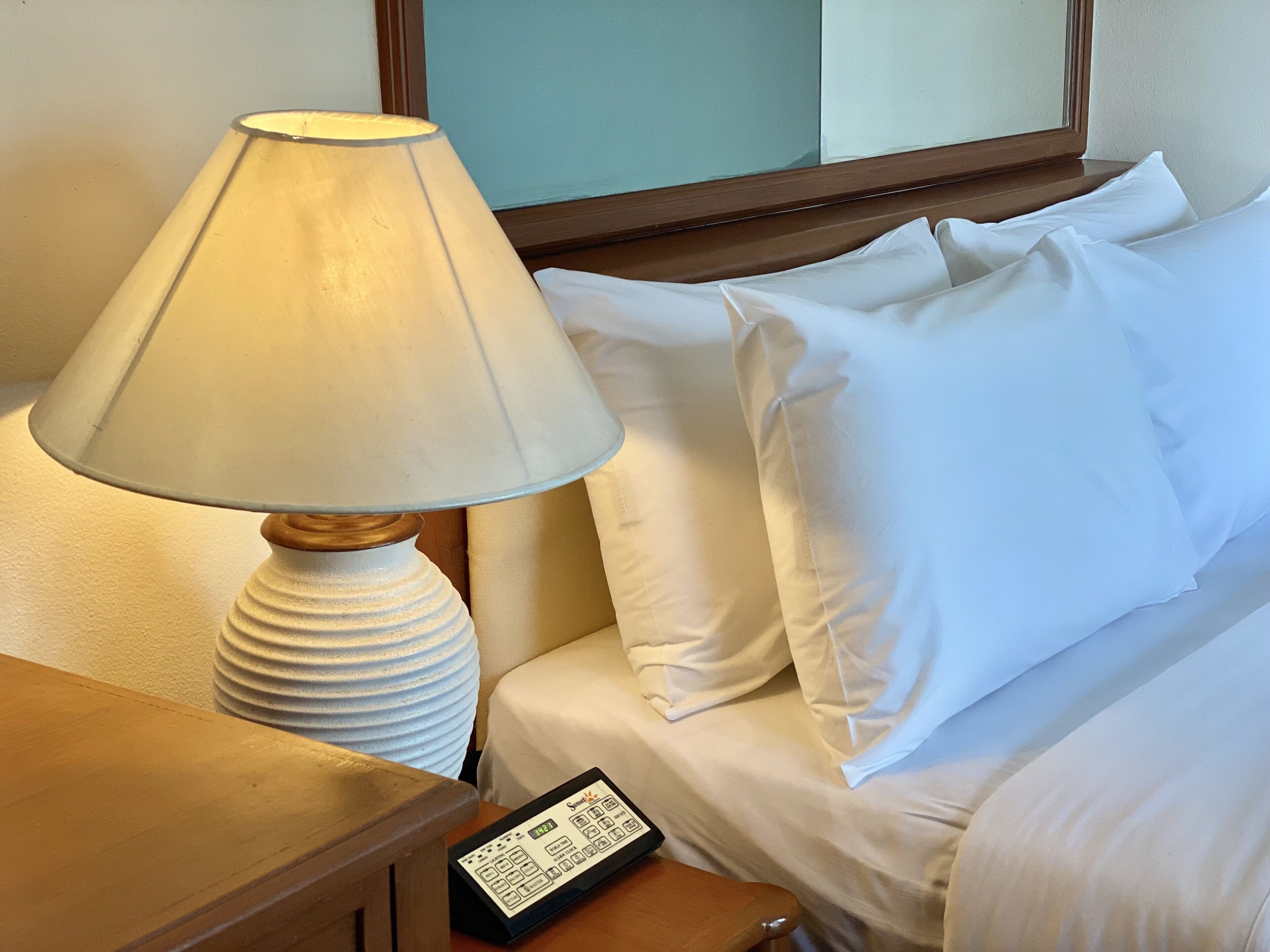 A hotel room with a bed, white pillows, a lamp, and a remote control on a wooden nightstand.