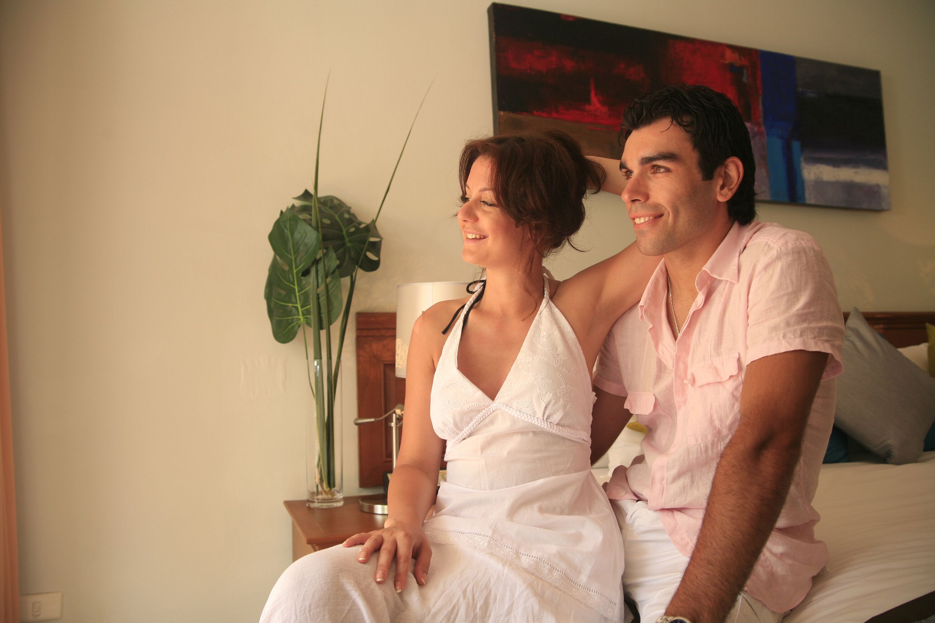 A couple sitting on a bed in a room with a large plant and a painting on the wall.