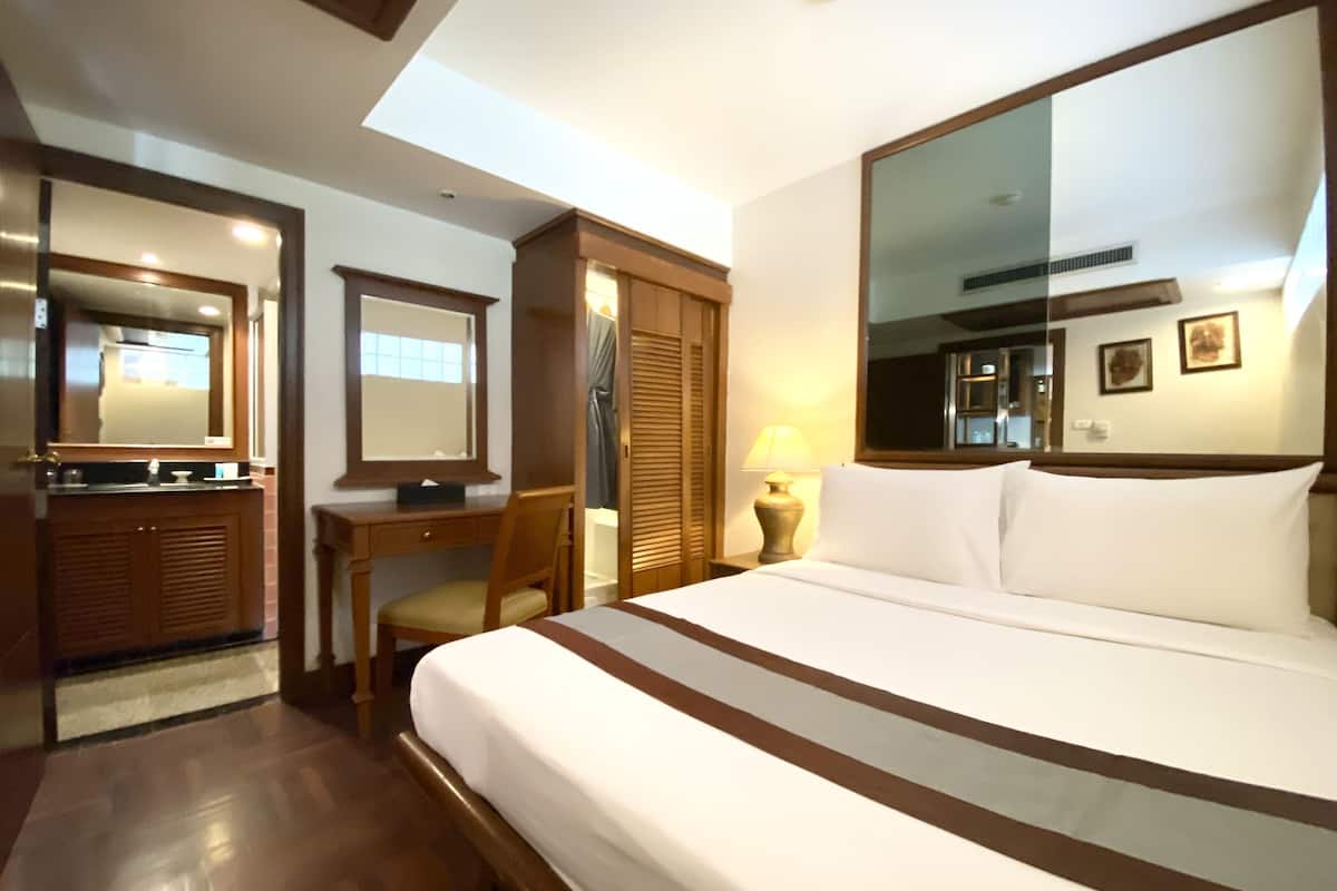 A hotel room with a large bed, a desk with a chair, a mirror, a wardrobe, and a bedside table with a lamp.