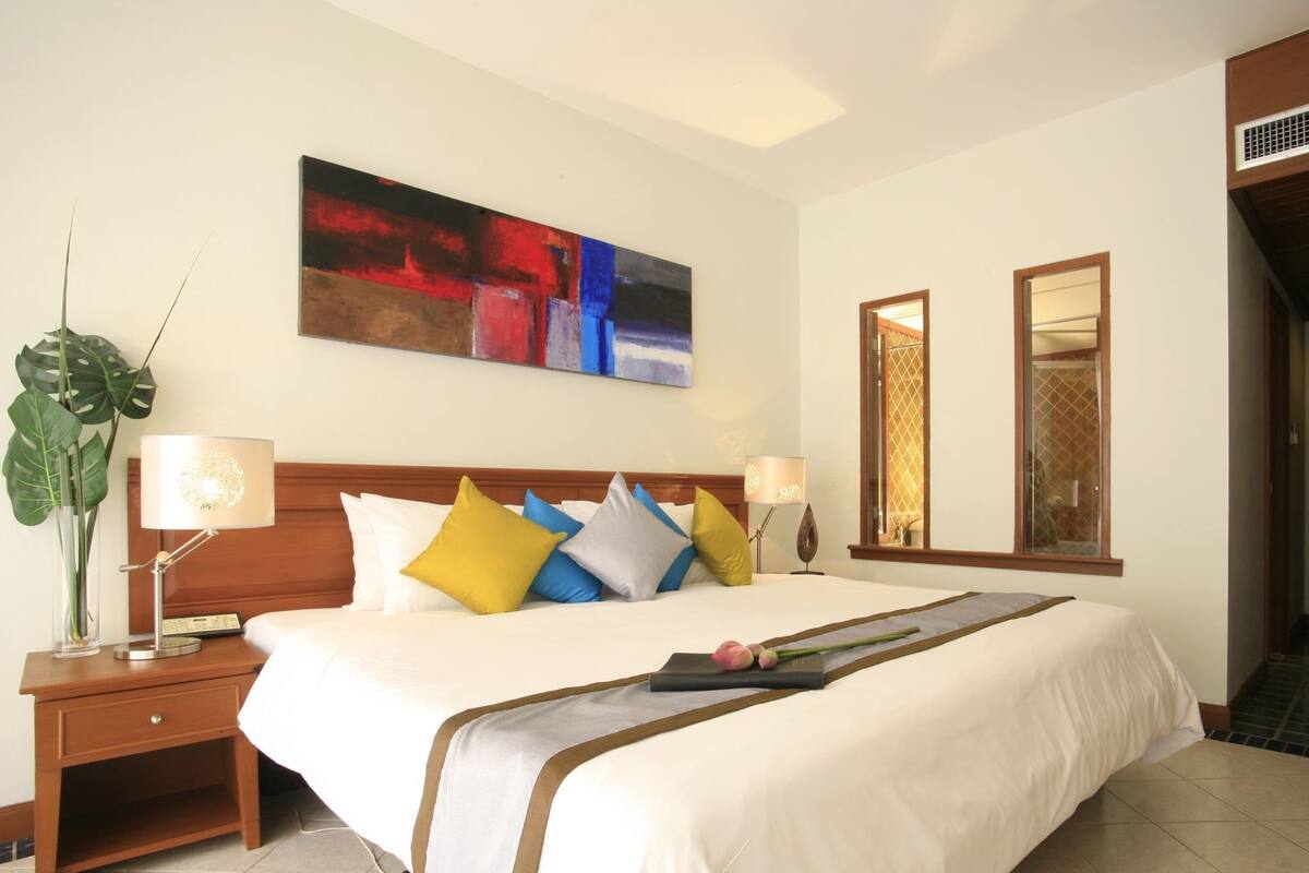 A hotel room with a large bed, a wooden headboard, a bedside table with a lamp, a large abstract painting on the wall, and a mirror above the bed.