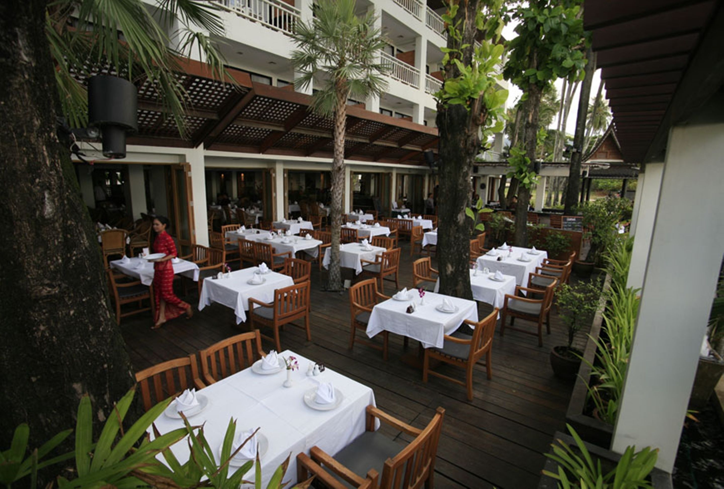 An outdoor dining area with tables and chairs, surrounded by trees and a building with balconies.