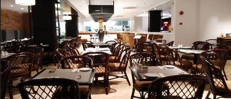 Un restaurante con varias mesas y sillas de mimbre, una zona de cocina al fondo y una decoración interior moderna.