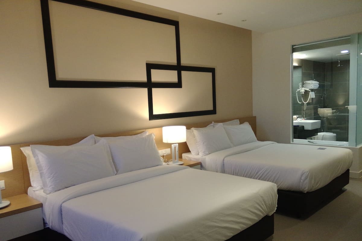 A hotel room with two beds, a large mirror, and a bathroom visible through the glass partition.
