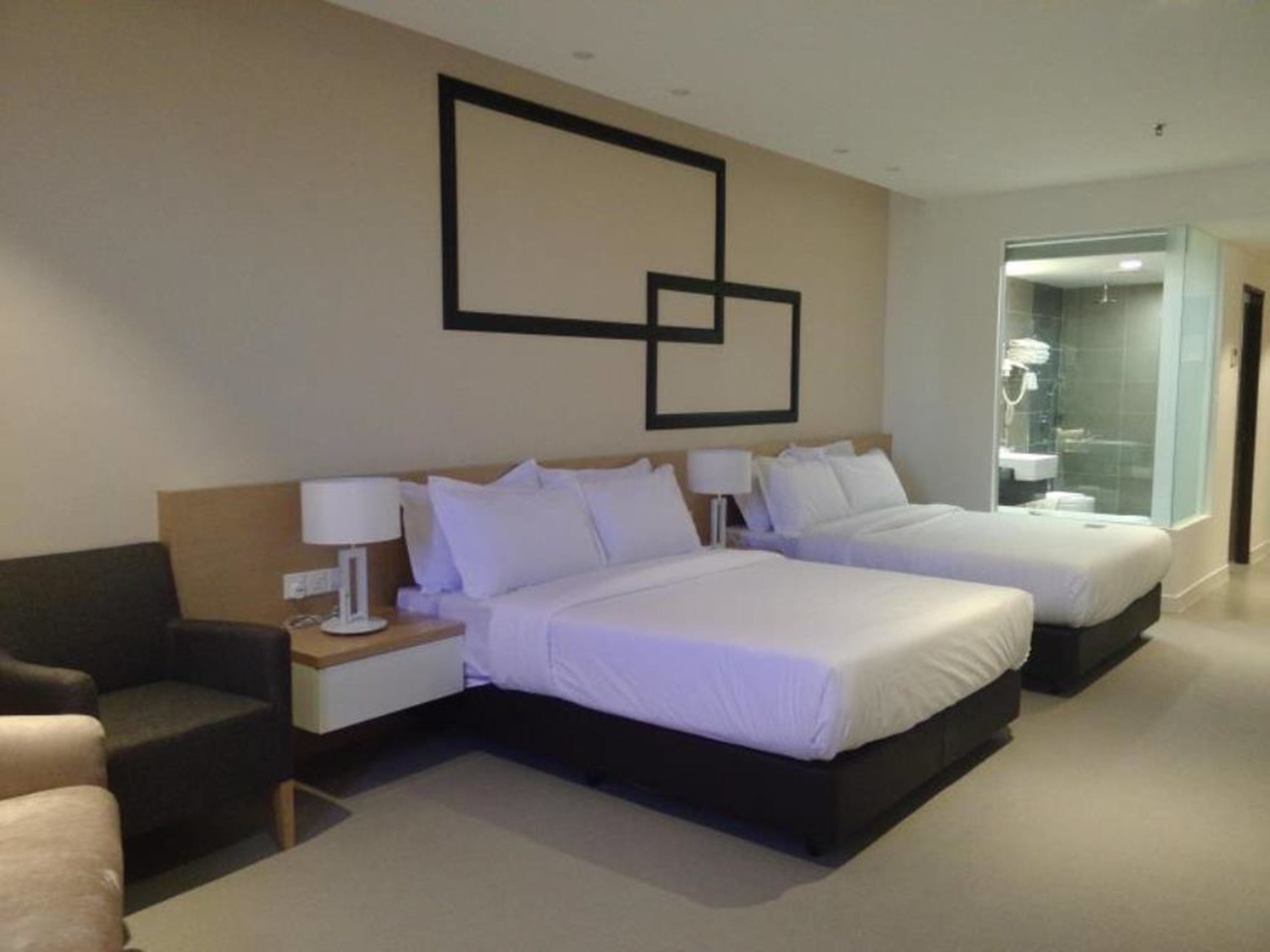 A modern hotel room with two beds, a sofa, a nightstand, and a bathroom visible through a glass door.
