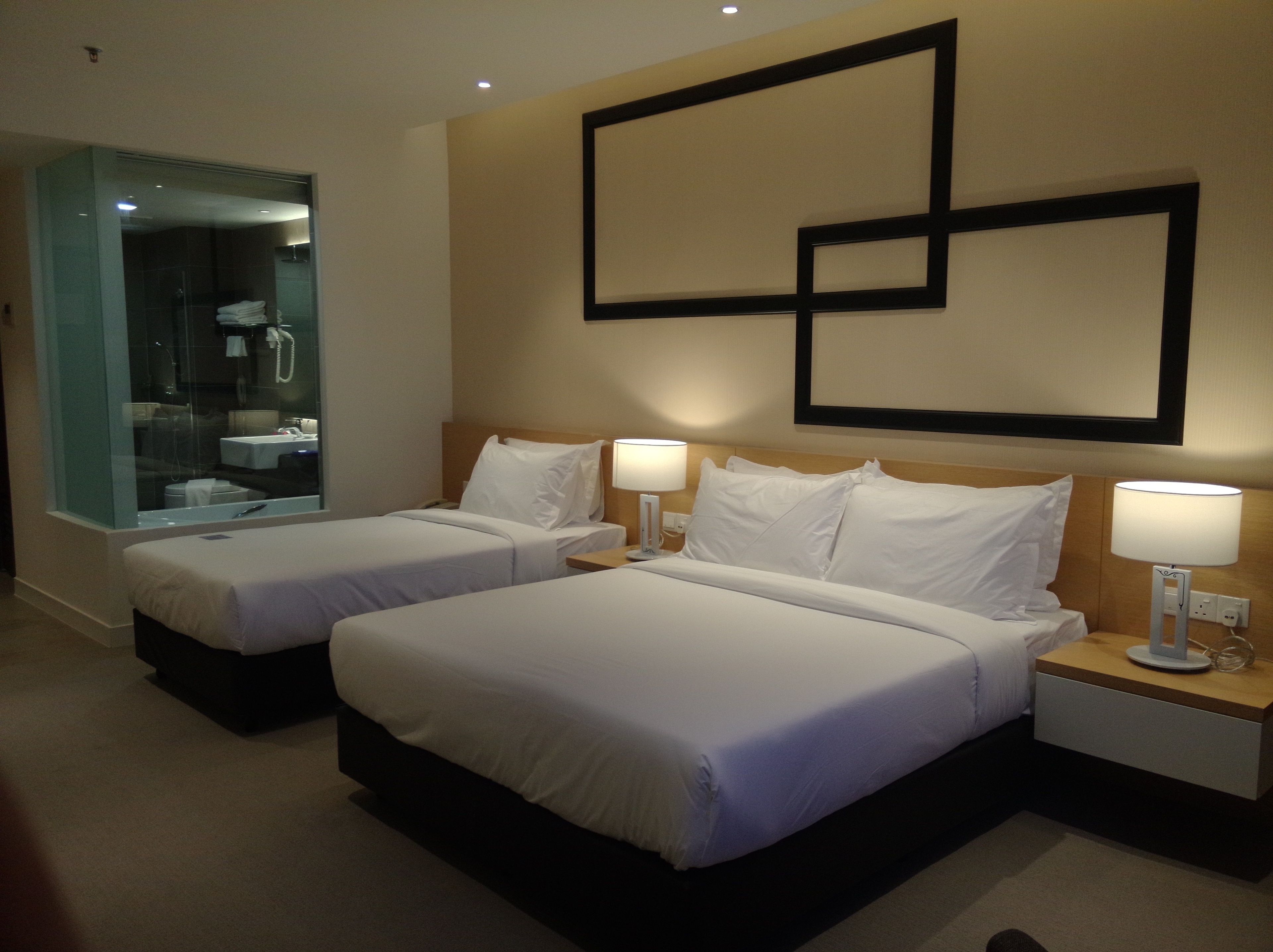 A hotel room with two beds, a bathroom with a glass partition, and a wall-mounted artwork.