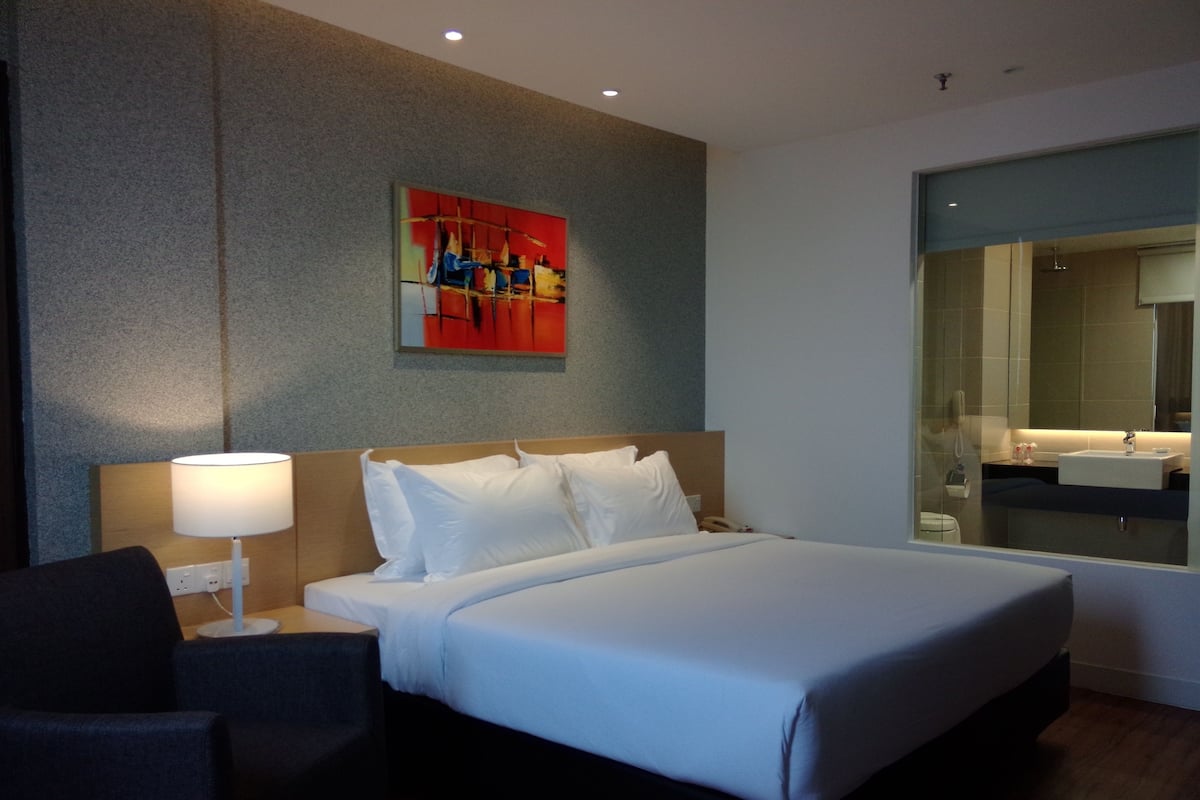 A hotel room with a bed, a bedside lamp, a chair, a painting on the wall, and a bathroom visible through a glass door.