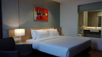Una habitación de hotel con cama, una lámpara de noche, una silla, un cuadro en la pared y un baño visible a través de una puerta de cristal.