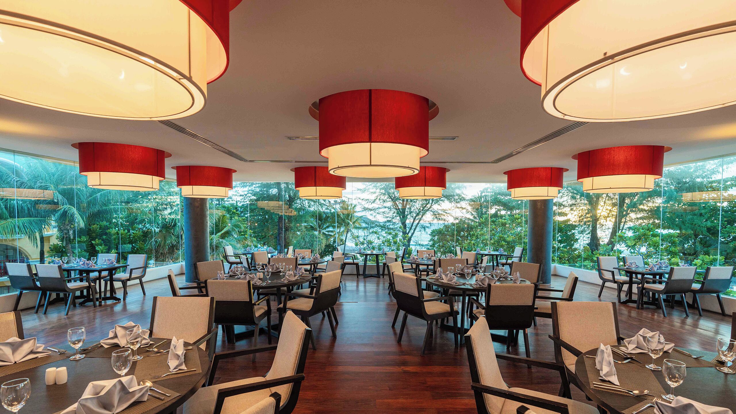 A restaurant with multiple tables and chairs, large red pendant lights, and a view of greenery outside.