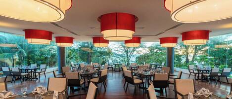 A restaurant with multiple tables and chairs, large red pendant lights, and a view of greenery outside.