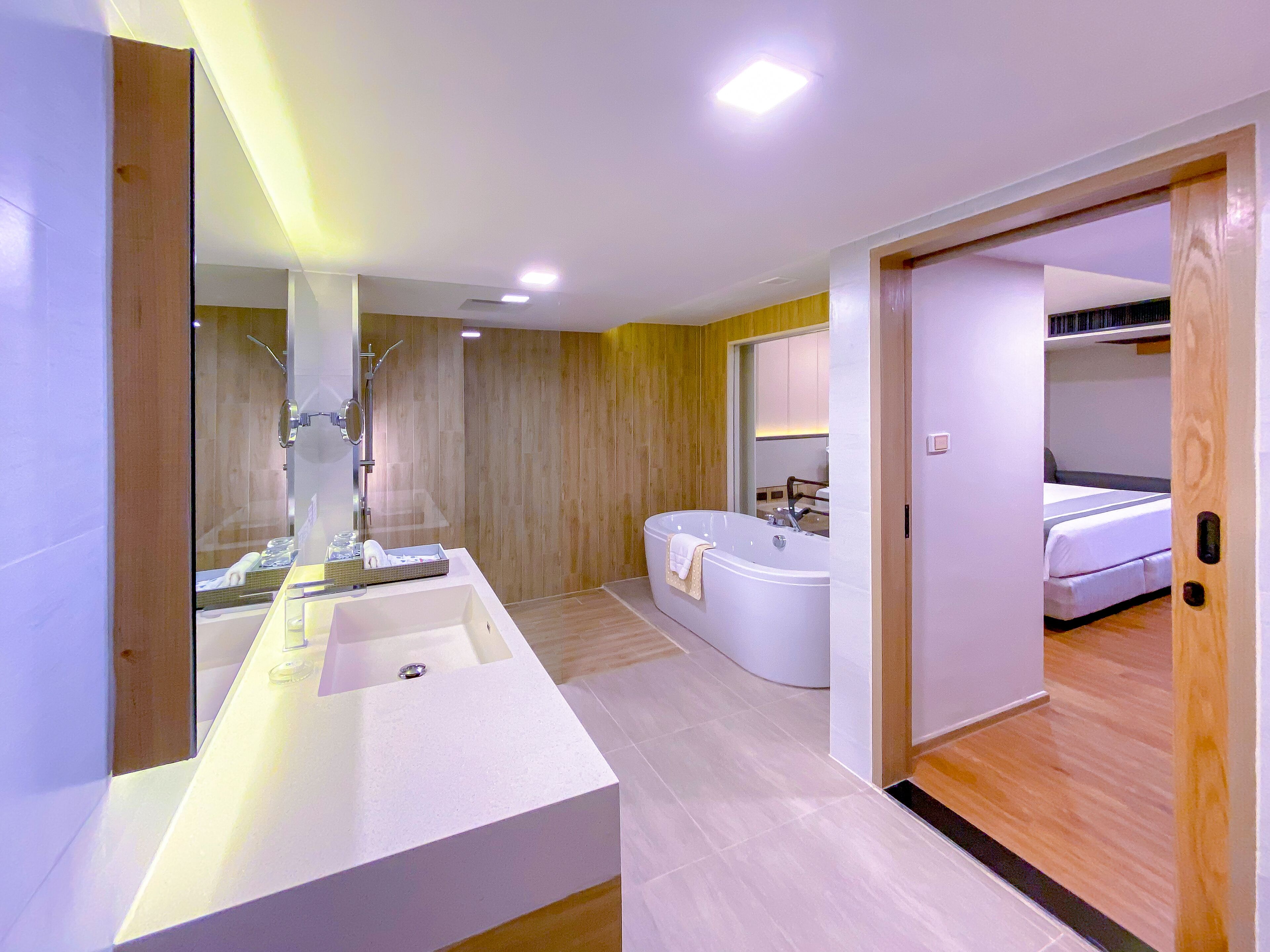 Ein modernes Badezimmer mit einem großen weißen Waschbecken, einer Badewanne und einer Dusche. Ein Holzboden und eine sichtbare Tür, die zu einem Schlafzimmer führt, sind ebenfalls vorhanden.