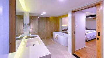 Ein modernes Badezimmer mit einem großen weißen Waschbecken, einer Badewanne und einer Dusche. Ein Holzboden und eine sichtbare Tür, die zu einem Schlafzimmer führt, sind ebenfalls vorhanden.