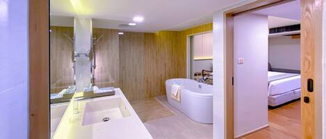 Un baño moderno con un lavamanos grande de color blanco, una bañera y una ducha. El suelo es de madera y se ve una puerta que da a un dormitorio.