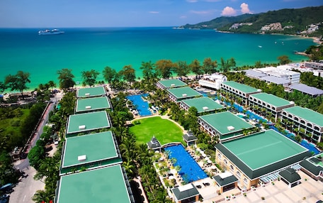 A resort with multiple green-roofed buildings, a central swimming pool, and a view of the ocean and hills.