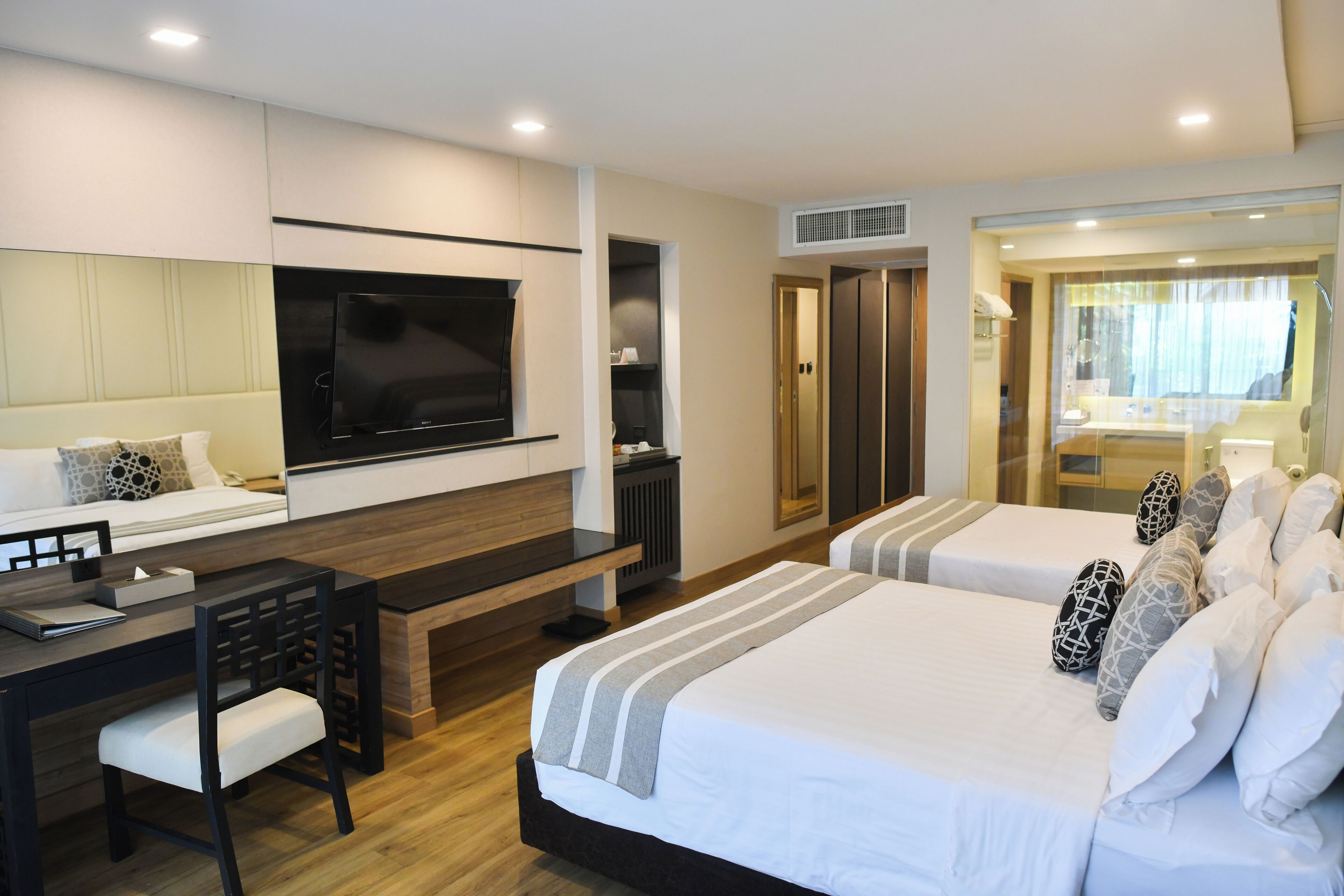 A modern hotel room with a large flat-screen TV, a desk with a chair, two beds with white bedding and patterned pillows, and a walk-in closet.