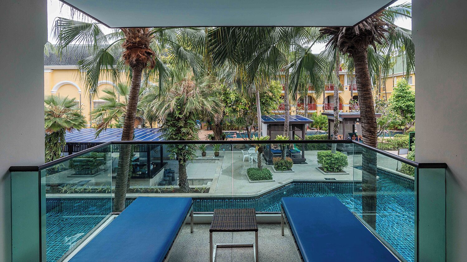 A poolside area with lounge chairs and a glass balustrade overlooking a pool and a courtyard with palm trees and buildings.