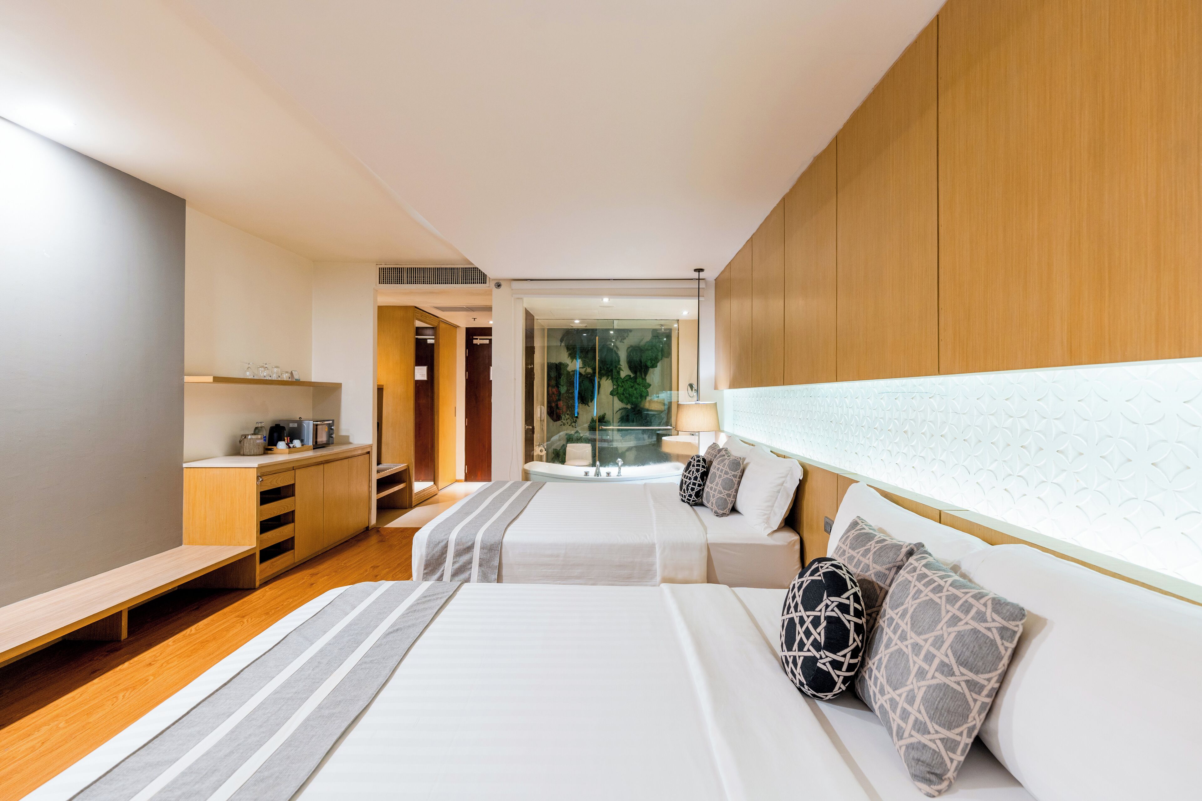 A modern hotel room with two beds, a wooden wardrobe, a small kitchenette, and a glass-enclosed bathroom.