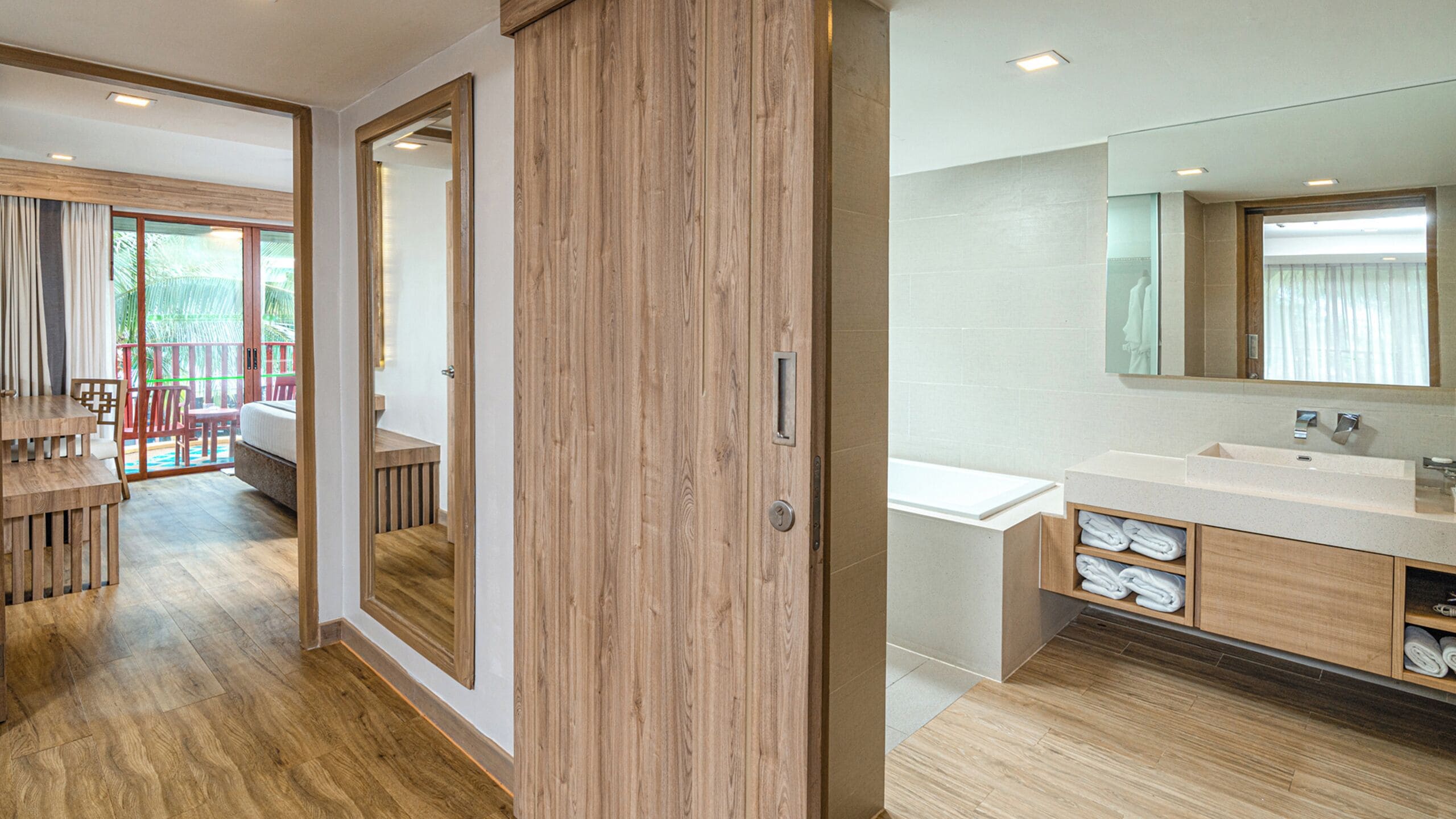 A modern hotel room with a wooden floor, a balcony with red chairs, a large mirror, and a bathroom with a sink and towel storage.