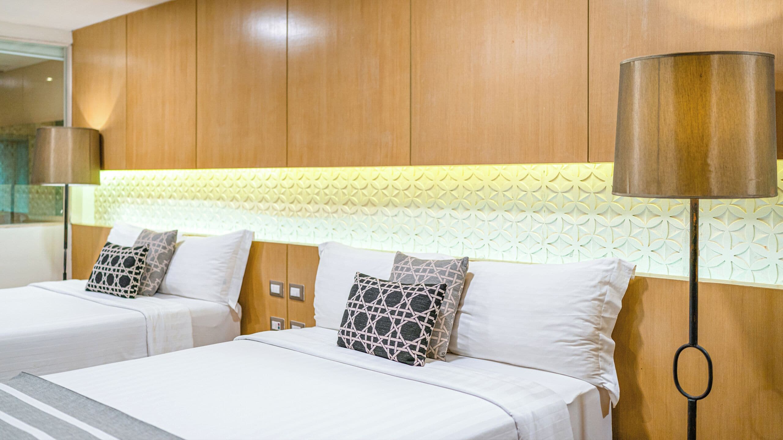 A hotel room with two beds, a wooden headboard, a decorative light panel, and a floor lamp.