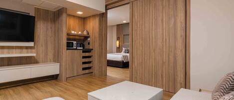 A modern hotel room with a flat-screen TV, a built-in kitchen with a microwave, a bed with a nightstand, and a large sliding door closet.