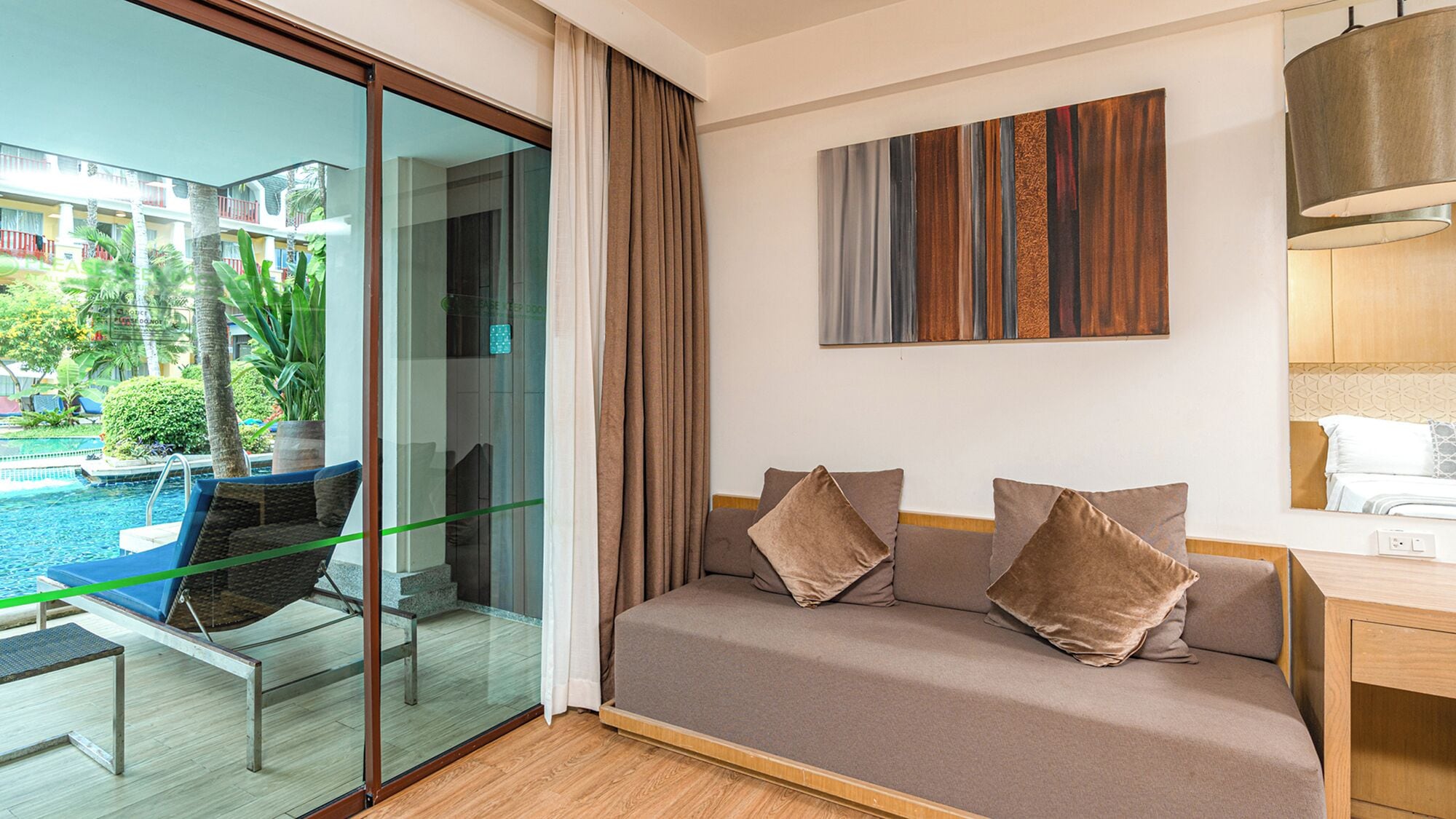 A modern hotel room with a glass door leading to a pool area, a sofa with cushions, and a wooden desk.