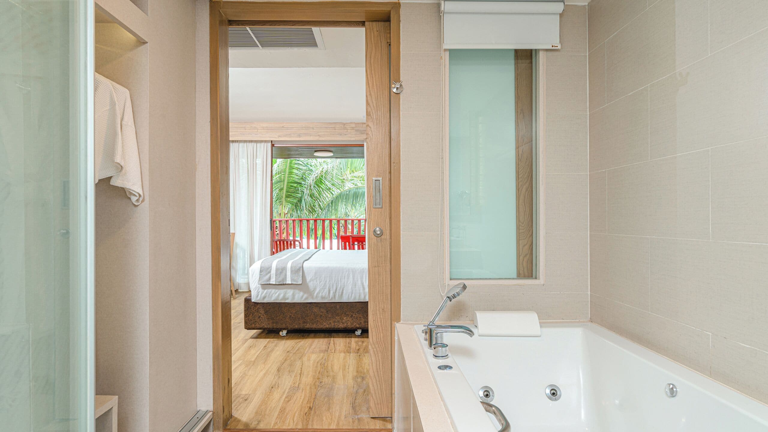 A modern bathroom with a large bathtub, a shower area, and a view of a balcony with a bed and red chairs.