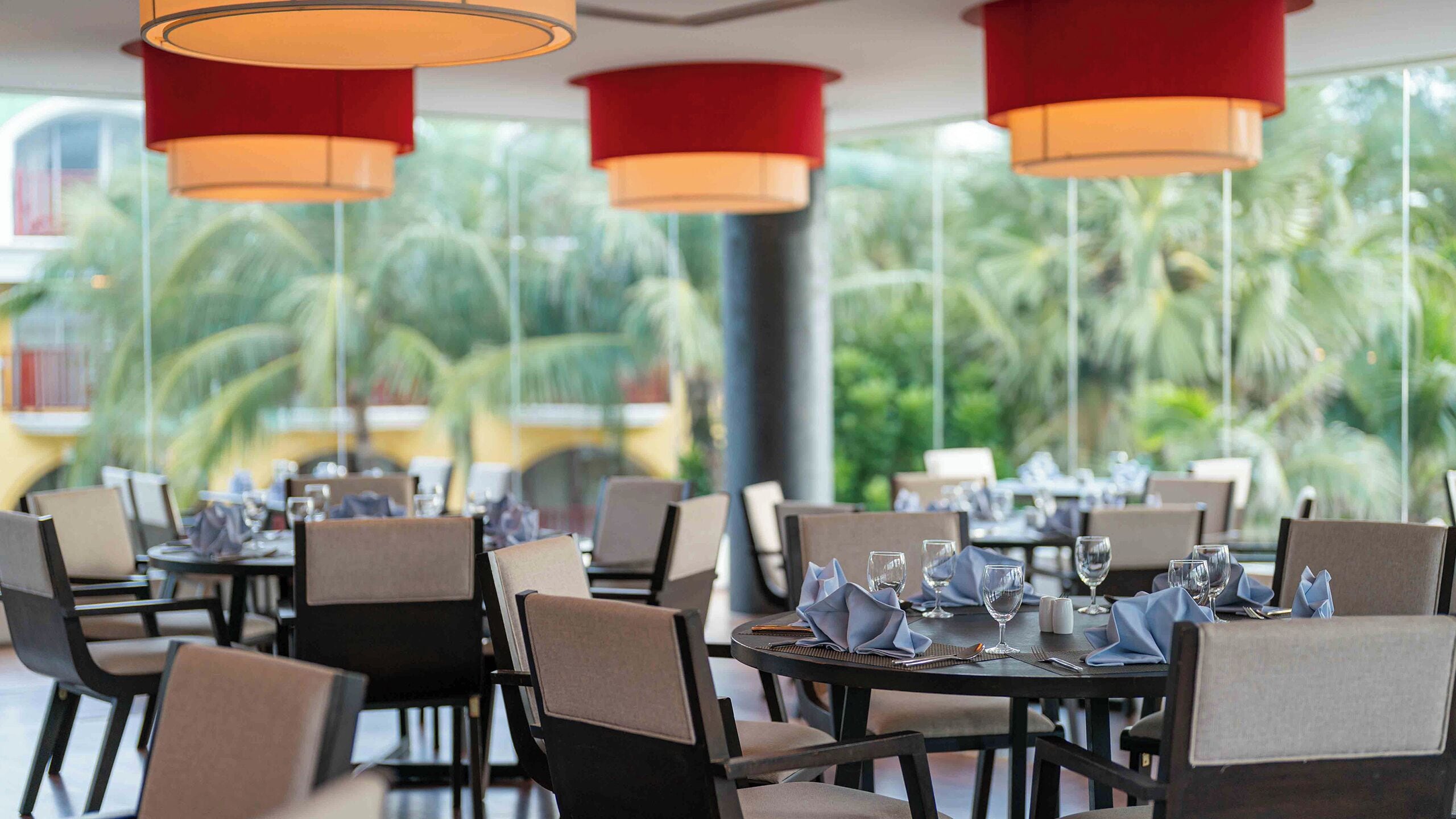 A restaurant with tables set for dining, featuring a tropical outdoor view with palm trees.