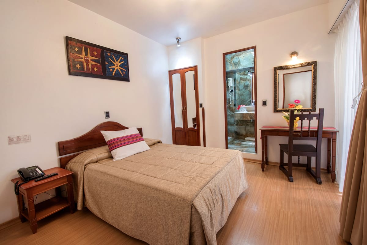 Un dormitorio con cama, mesita de noche, escritorio, silla, espejo y una obra de arte en la pared.
