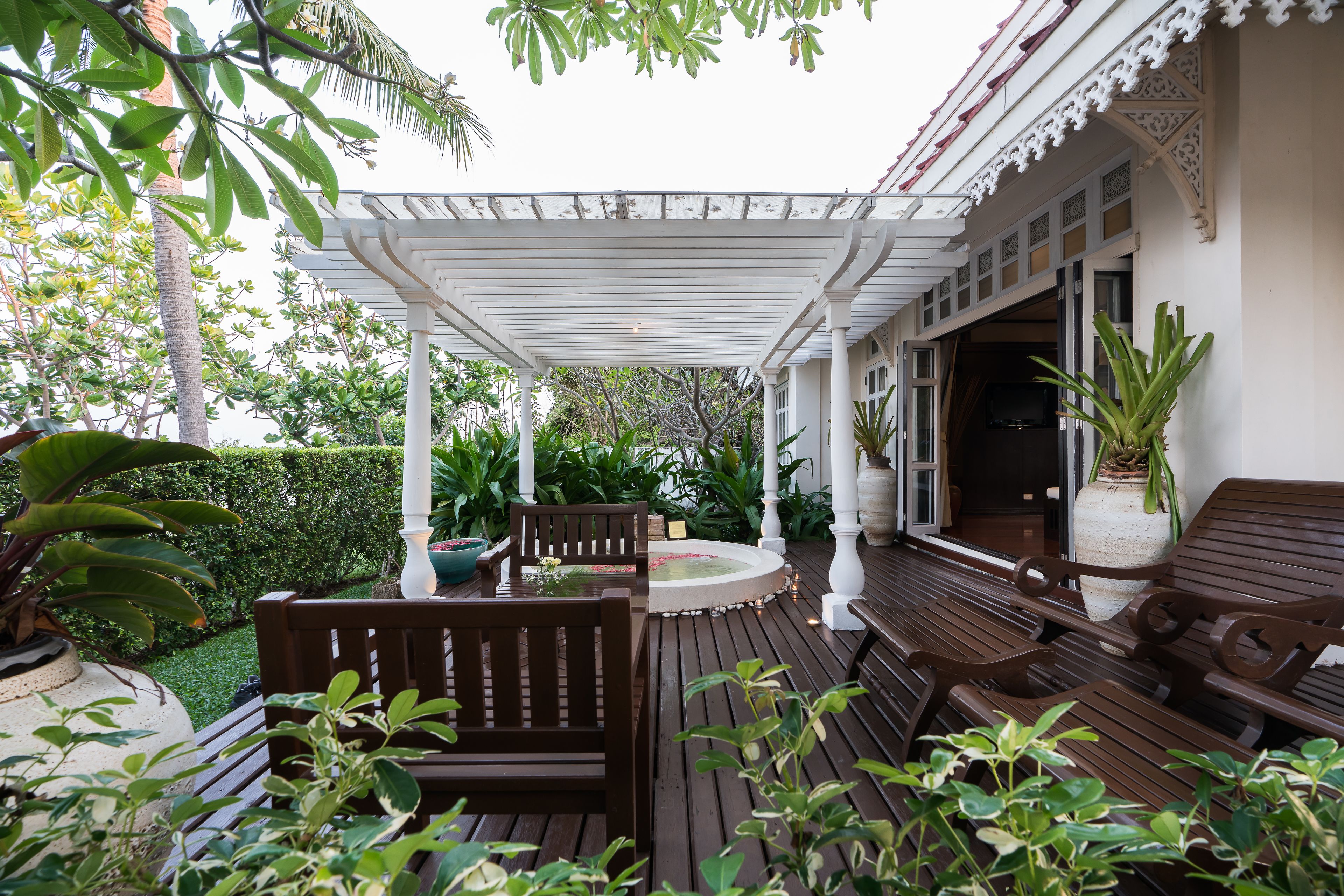 A wooden deck with benches and a small fountain, surrounded by lush greenery and a white pergola.