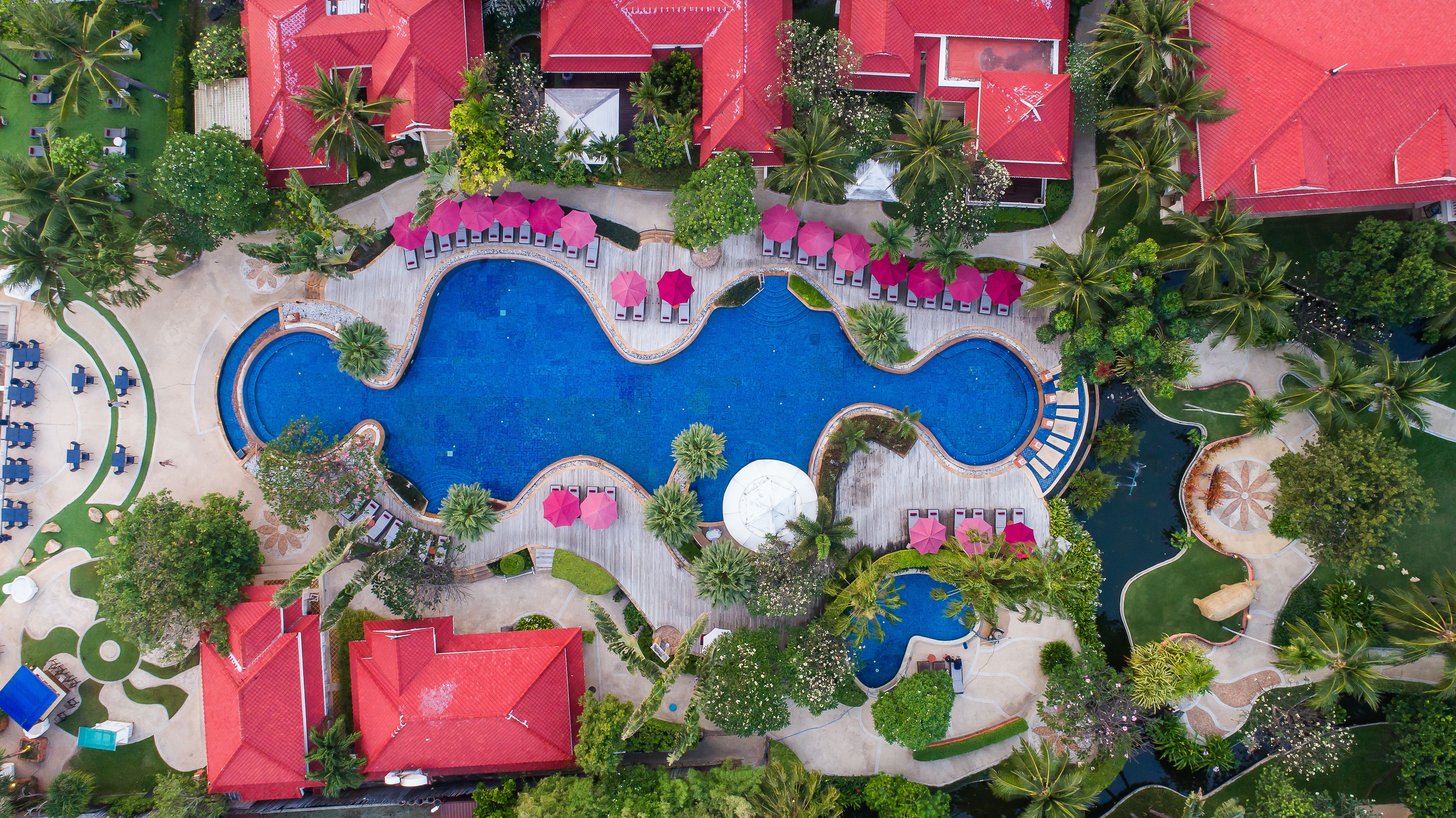 Luchtbeeld van een resort met een centraal zwembad, omringd door loungegedeeltes, palmbomen en gebouwen met rode daken.