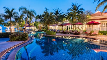 A swimming pool with lounge chairs and umbrellas, surrounded by palm trees and a building with a tiled roof.