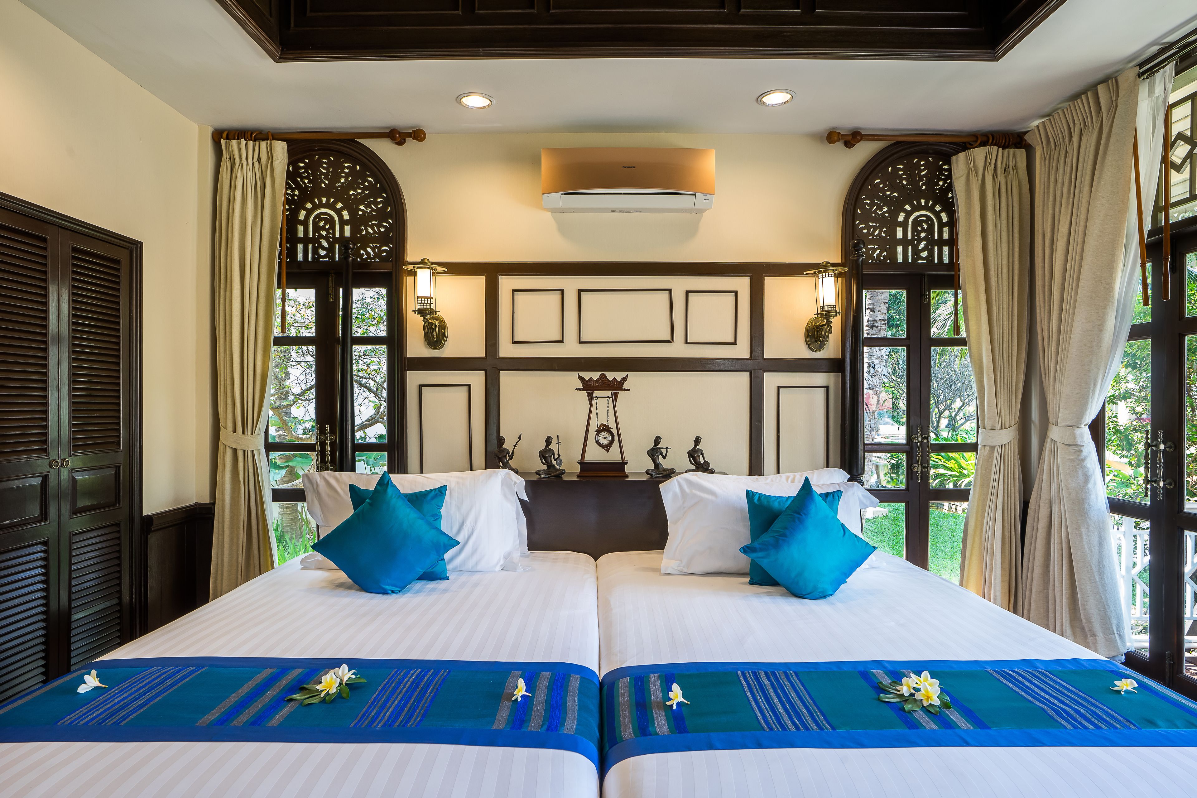 Cama doble con ropa de cama blanca y detalles azules, dos aires acondicionados de pared y vistas a zonas verdes a través de amplias ventanas.