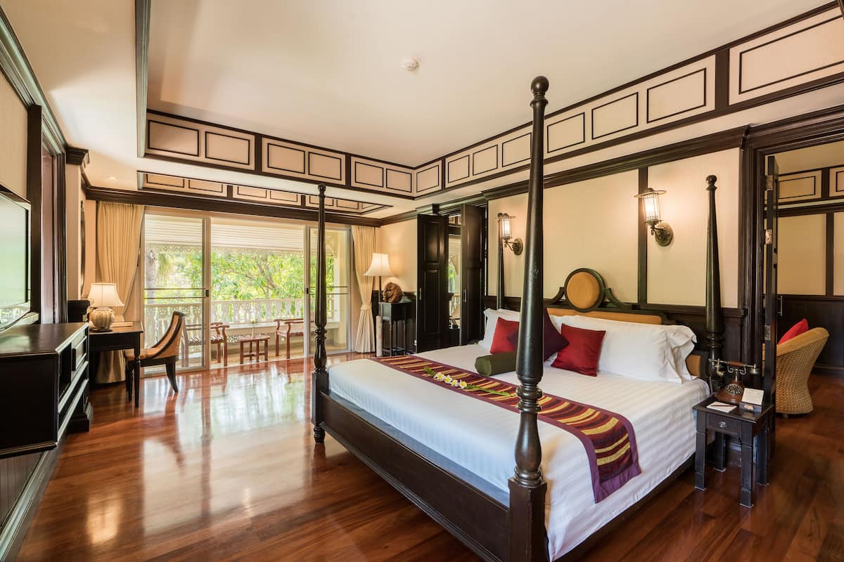 A four-poster bed with a wooden frame, a desk with a lamp, and a chair in a room with wooden flooring and large windows.